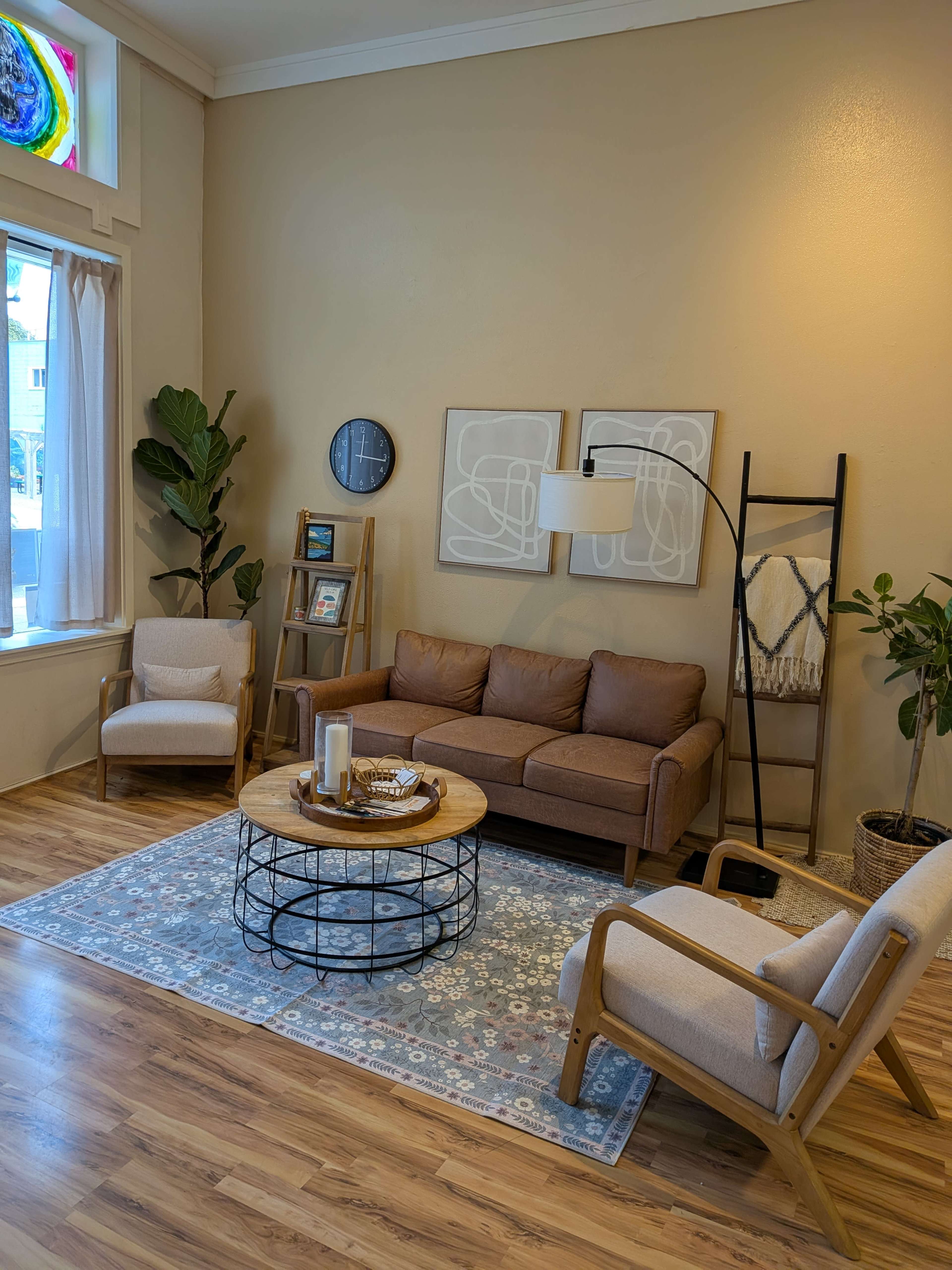 The image shows a cozy living room featuring a brown sofa, two light-colored armchairs, a round wooden coffee table, and abstract wall art, with green plants and a side ladder shelf in the decor.