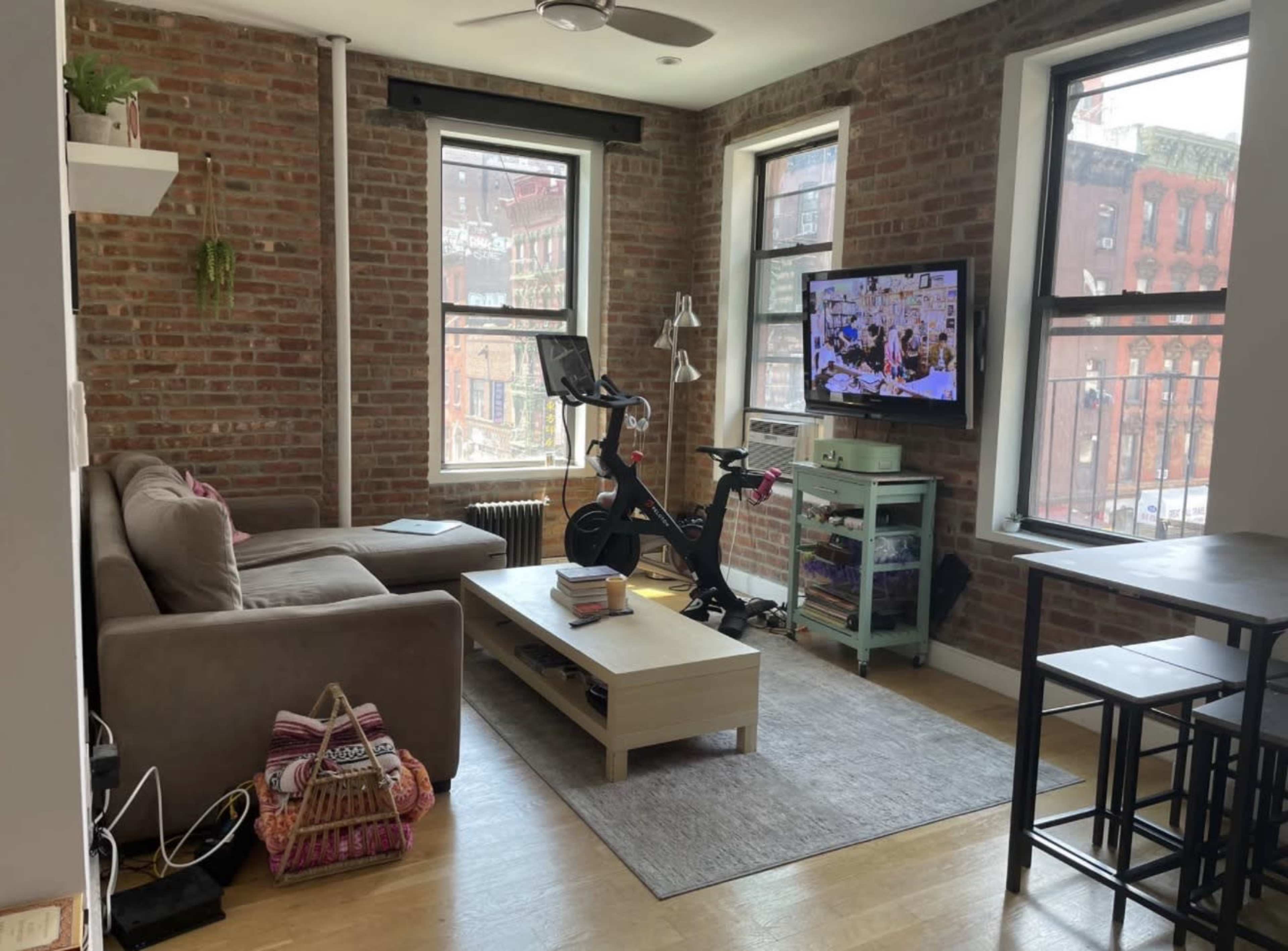 The image shows a compact living room with exposed brick walls, a sofa, a television, a stationary bike, and a small dining table.