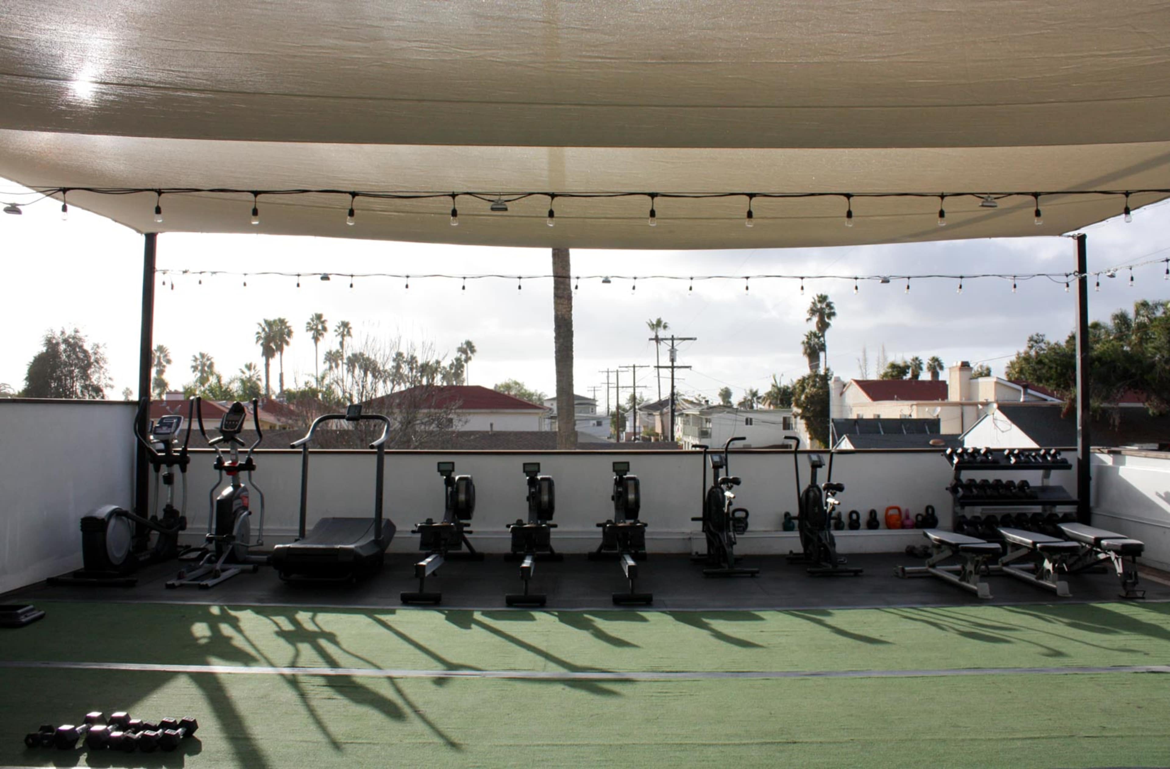 An outdoor fitness area with various exercise equipment, including treadmills, rowing machines, and free weights, under a shaded canopy.