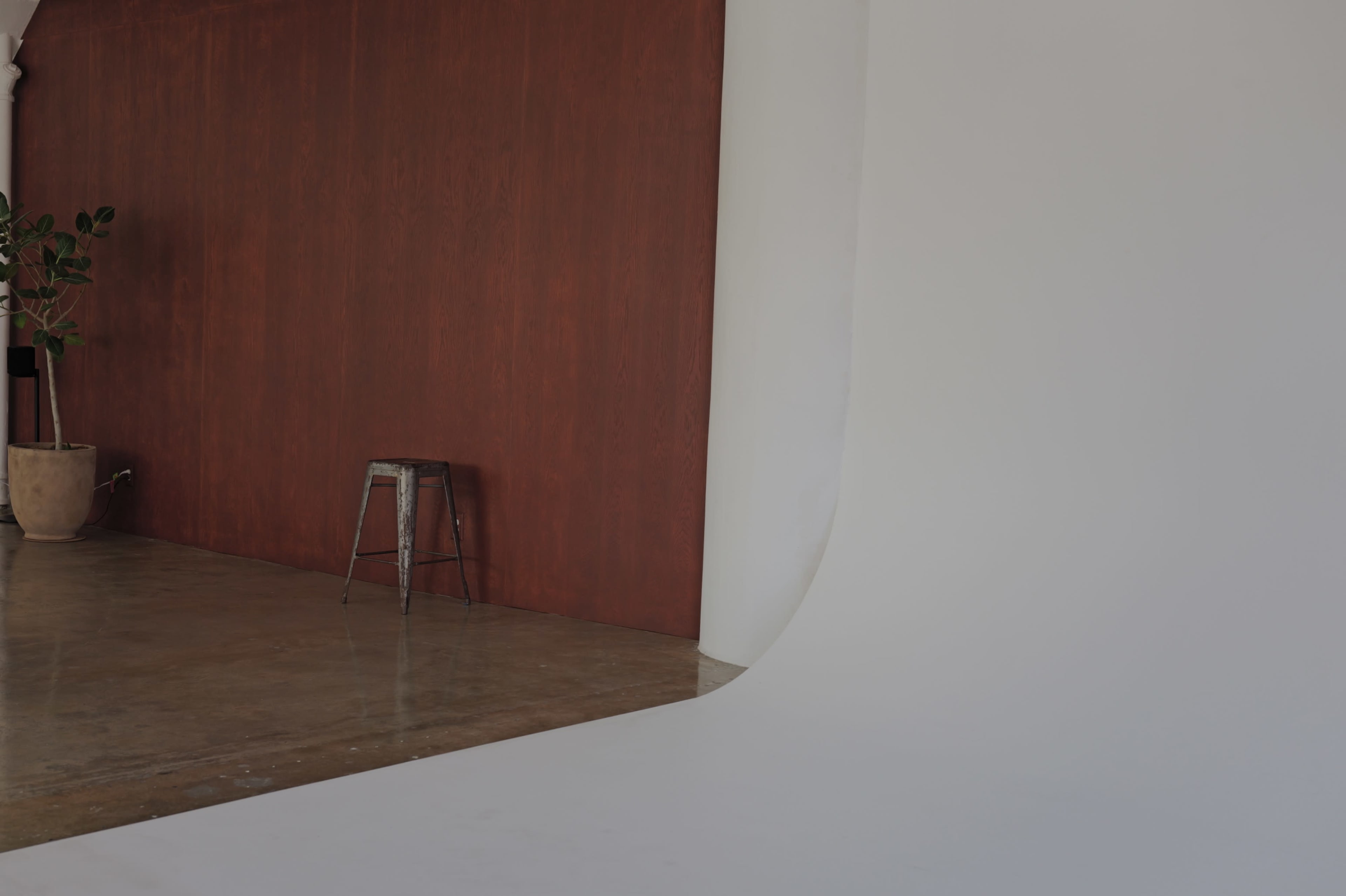 The image shows a photo studio with a wooden red wall, a white seamless backdrop, and a metal stool placed on a concrete floor.