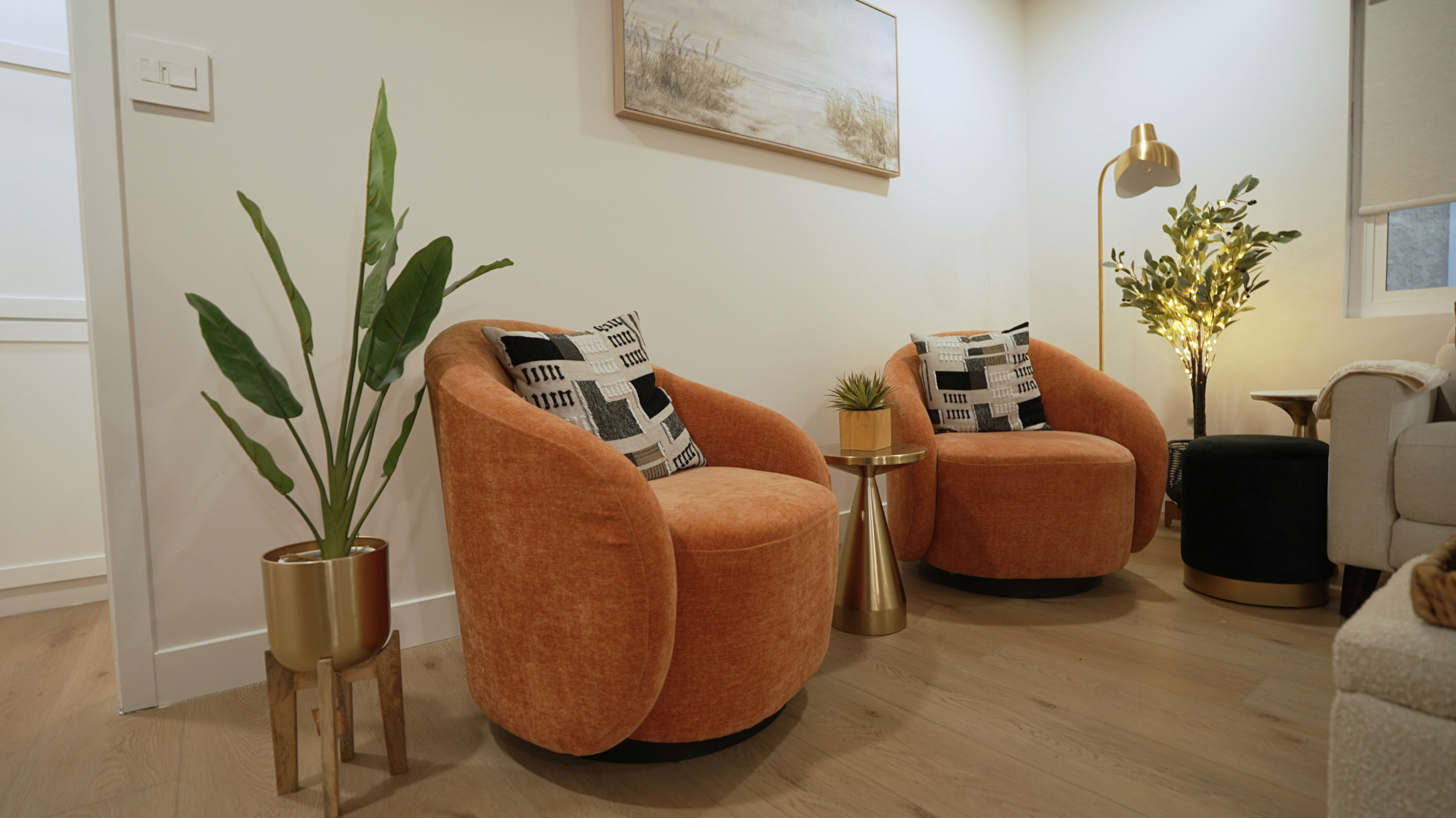 The image shows two orange armchairs with patterned cushions positioned beside a small gold side table and potted plants in a well-lit room.