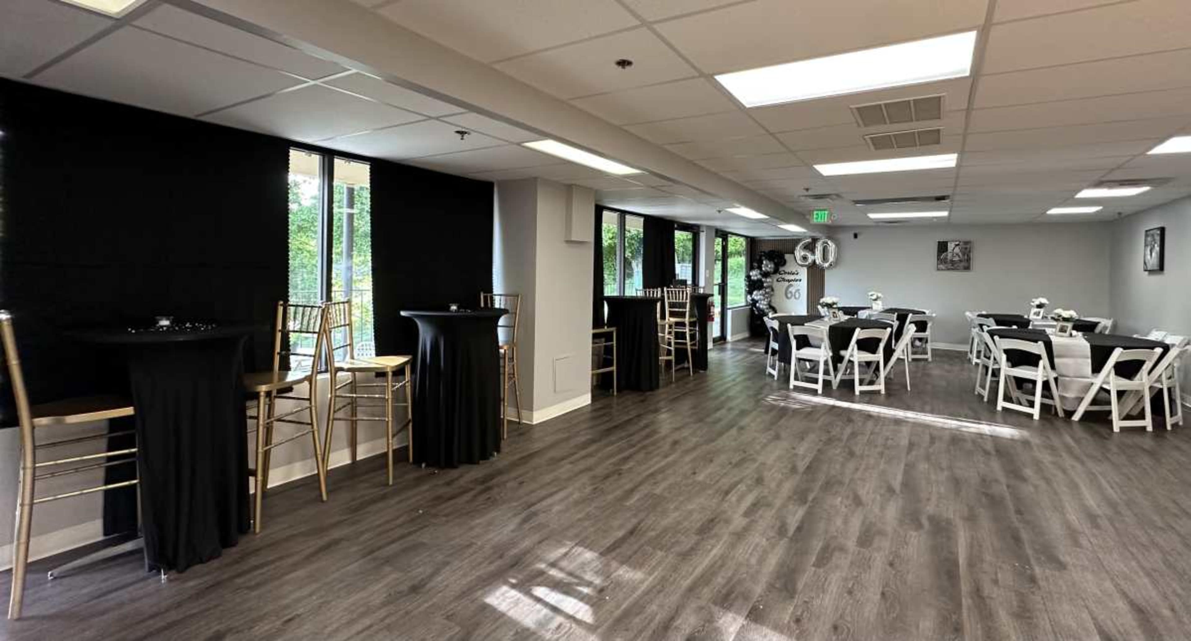 The image shows a spacious event room with black tablecloths on cocktail tables and white table settings arranged for dining, illuminated by natural light from large windows.