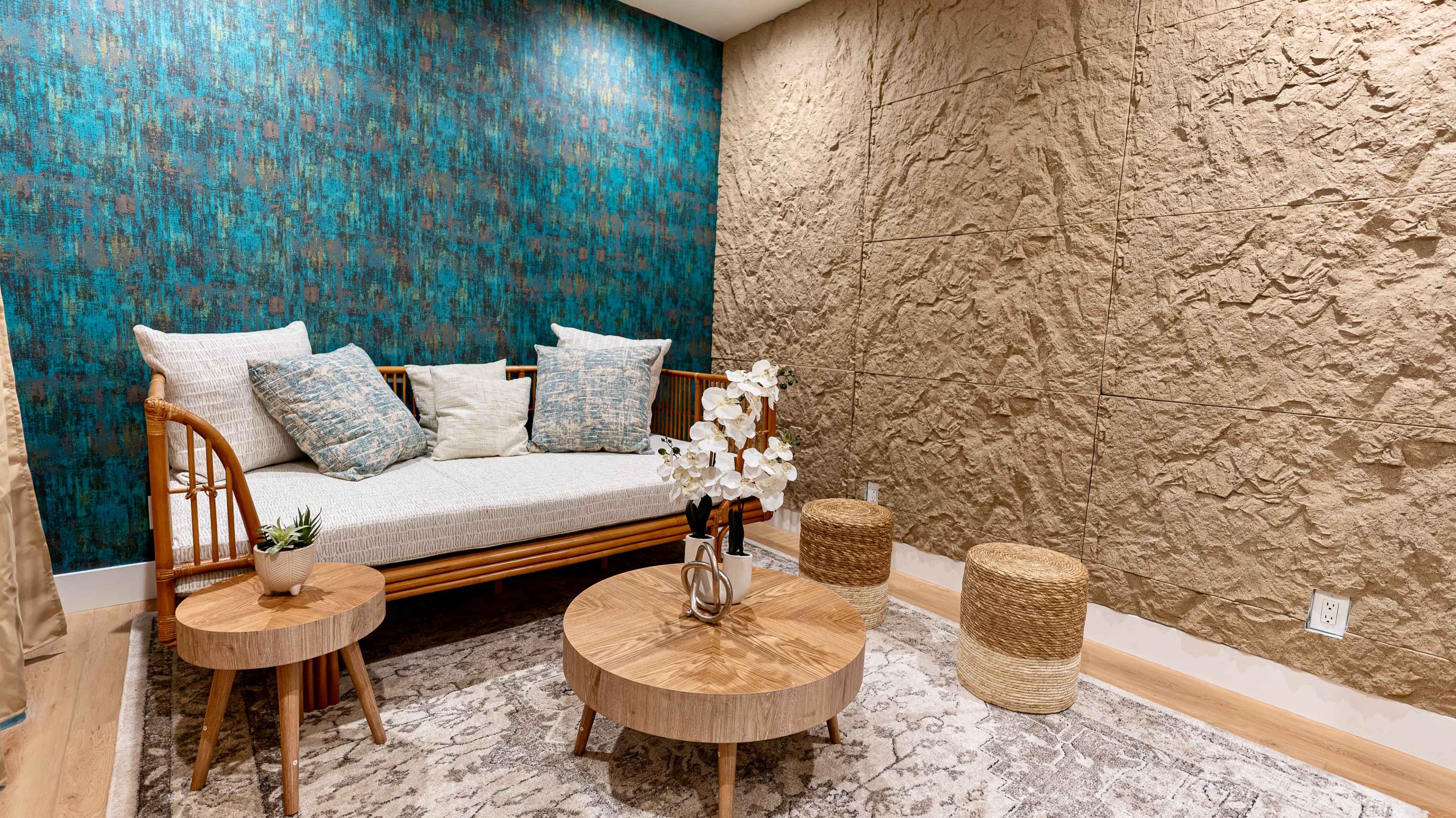 The room features a bamboo loveseat with cushions, a textured turquoise wall, natural fiber stools, and decorative plants on wooden tables.