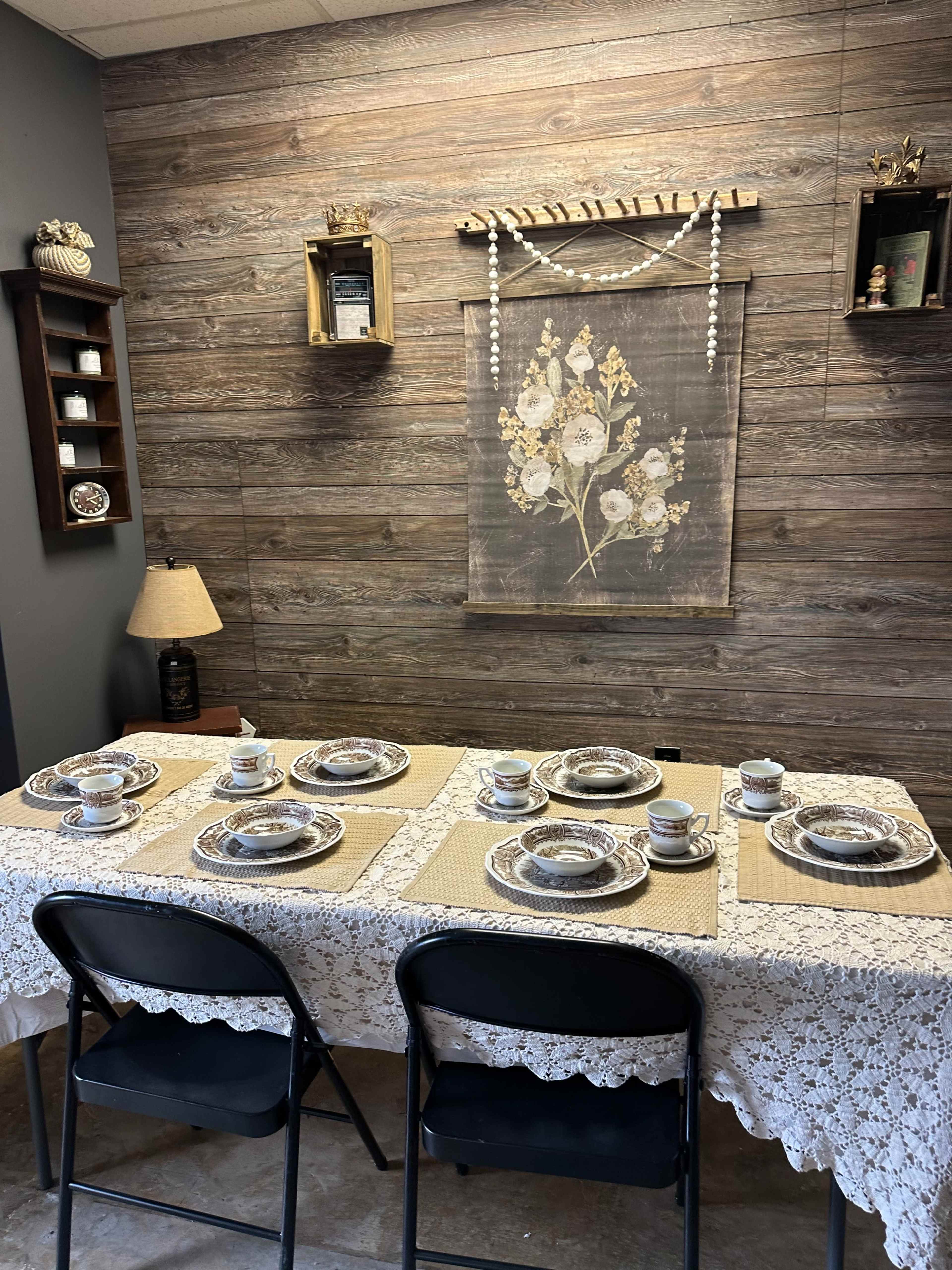 A dining table is set with eight place settings, featuring floral-patterned plates and cups, under decorative wall hangings in a room with wood-paneled walls.