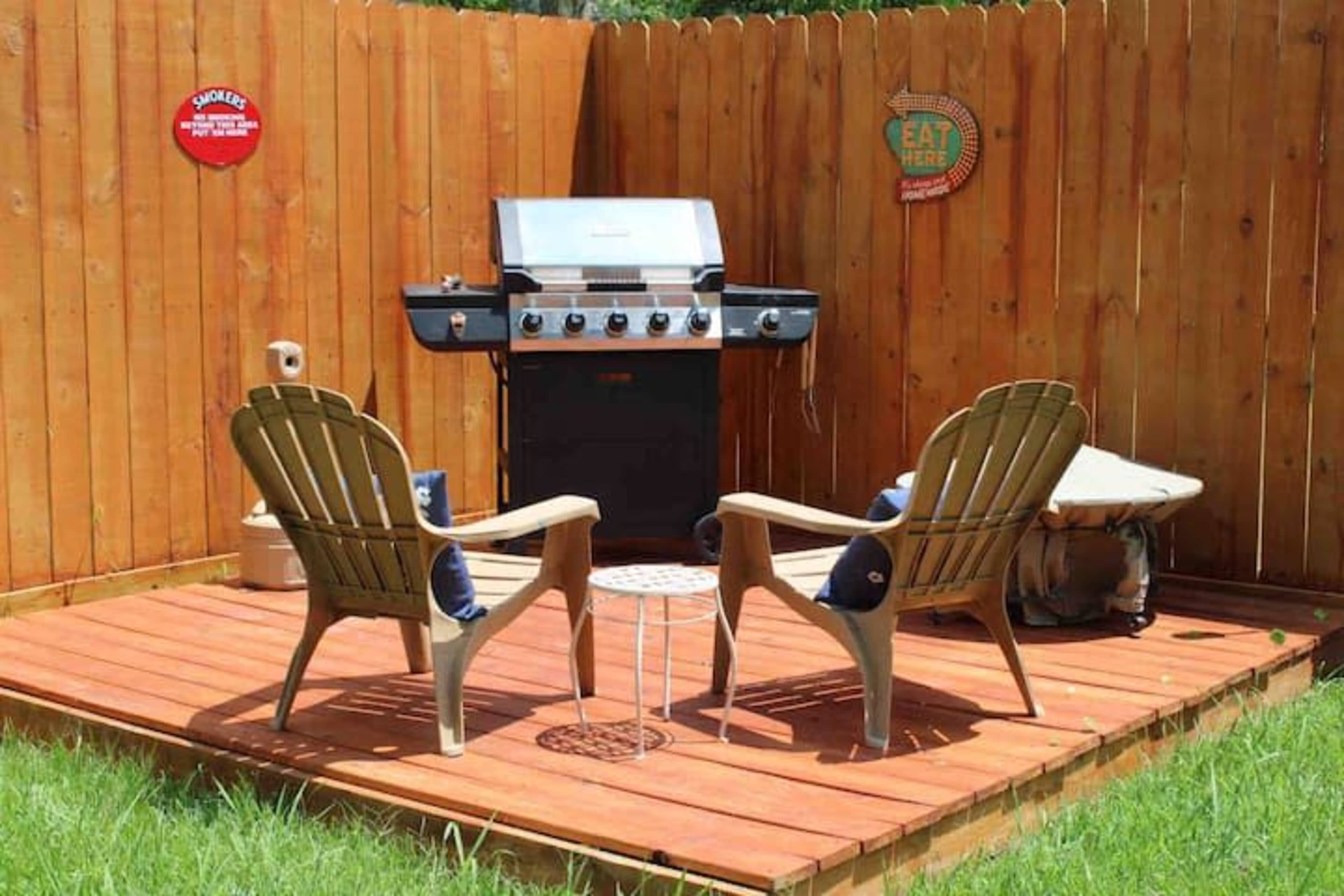 The image shows a wooden deck with a barbecue grill and two adirondack chairs surrounded by a wooden fence.