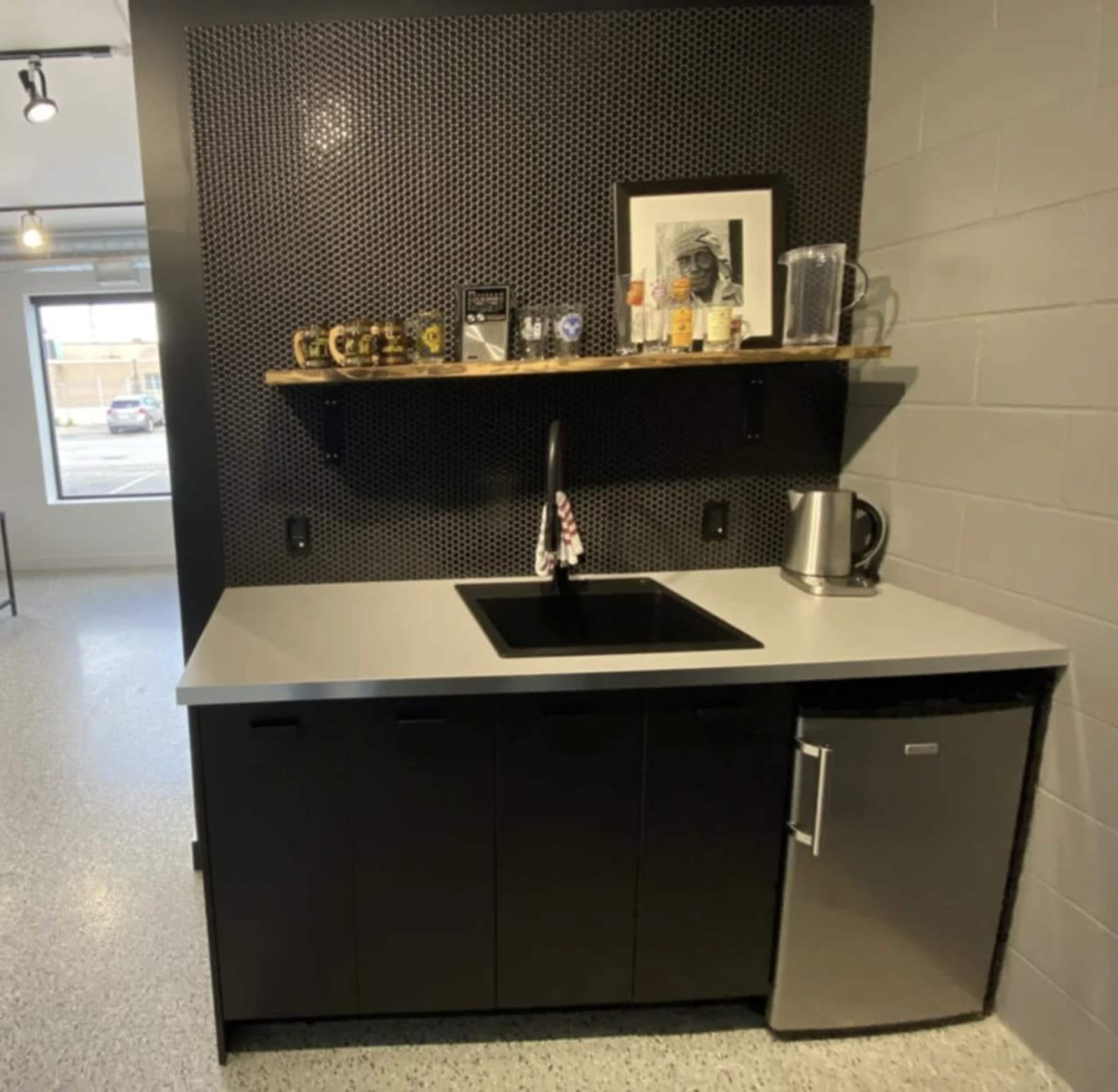 The image shows a modern kitchenette with a black countertop, a sink, a kettle, and a small refrigerator, all against a textured black wall and a wooden shelf displaying glassware and mugs.