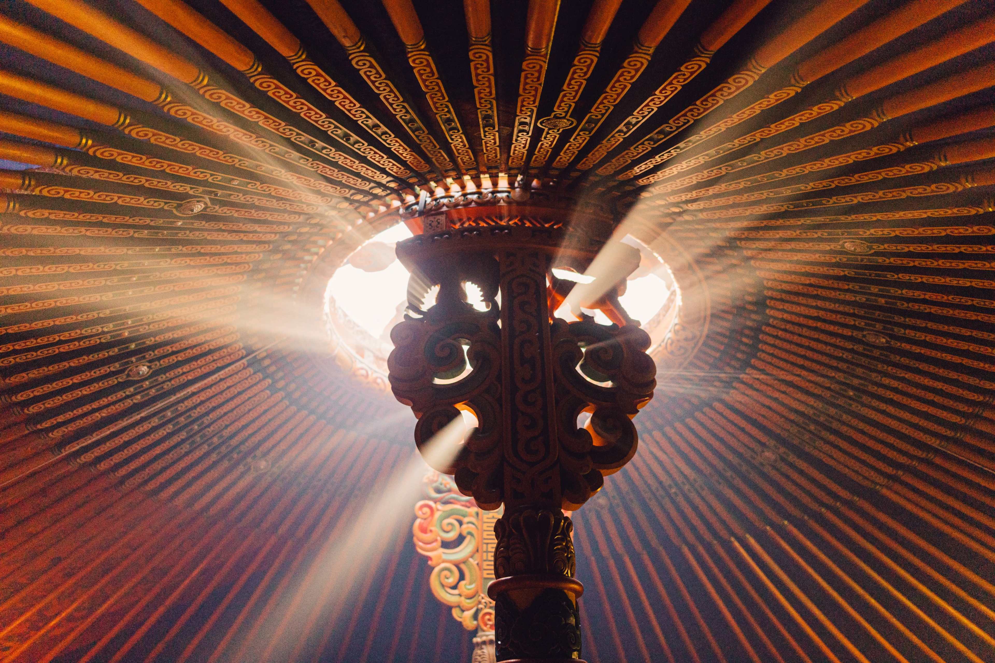Light streams through the circular opening at the top of a yurt, illuminating the intricate wooden structure and beams.
