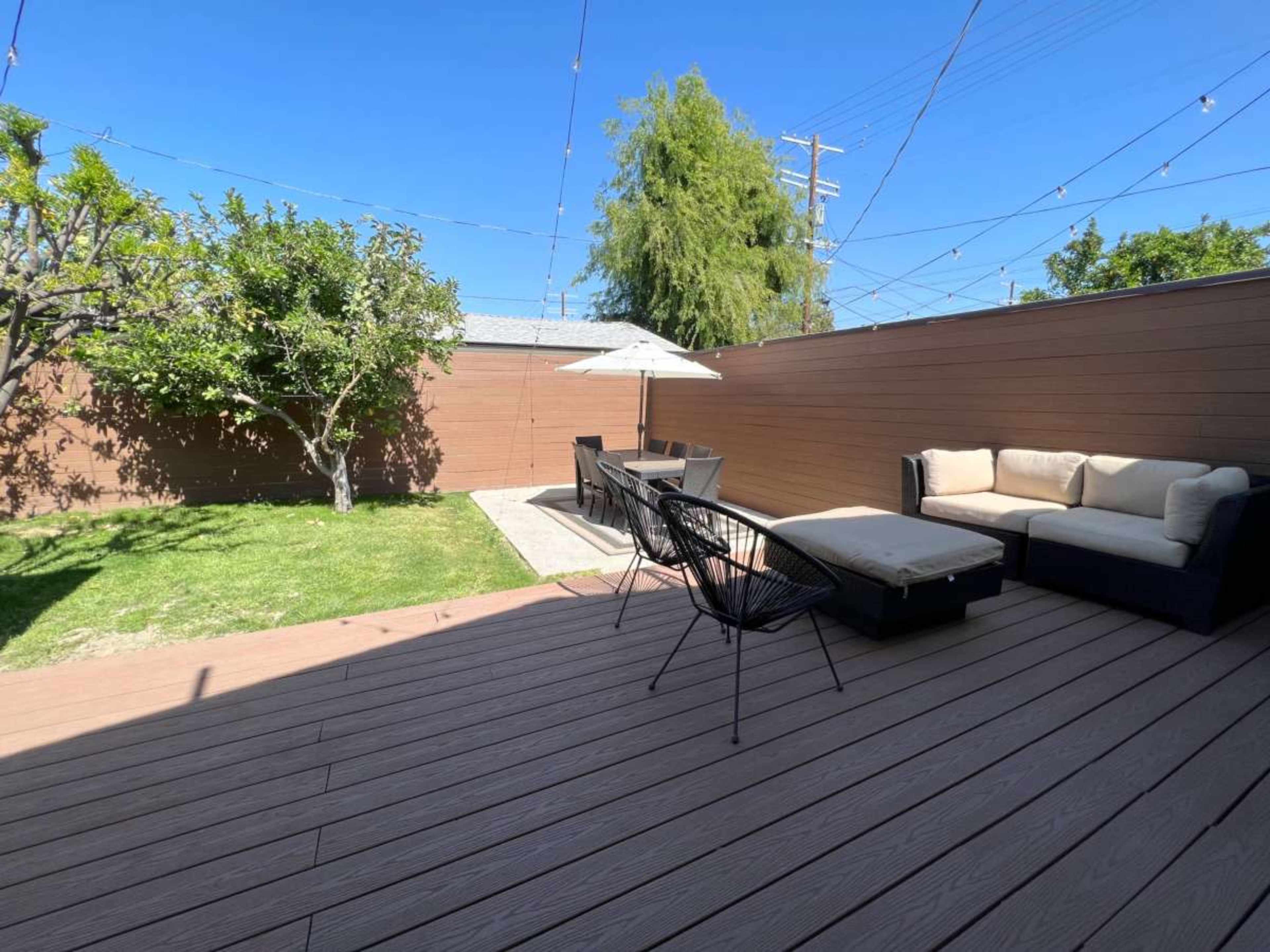 The image shows a backyard with a wooden deck featuring outdoor seating, a dining area, a lawn, and trees under a clear blue sky.