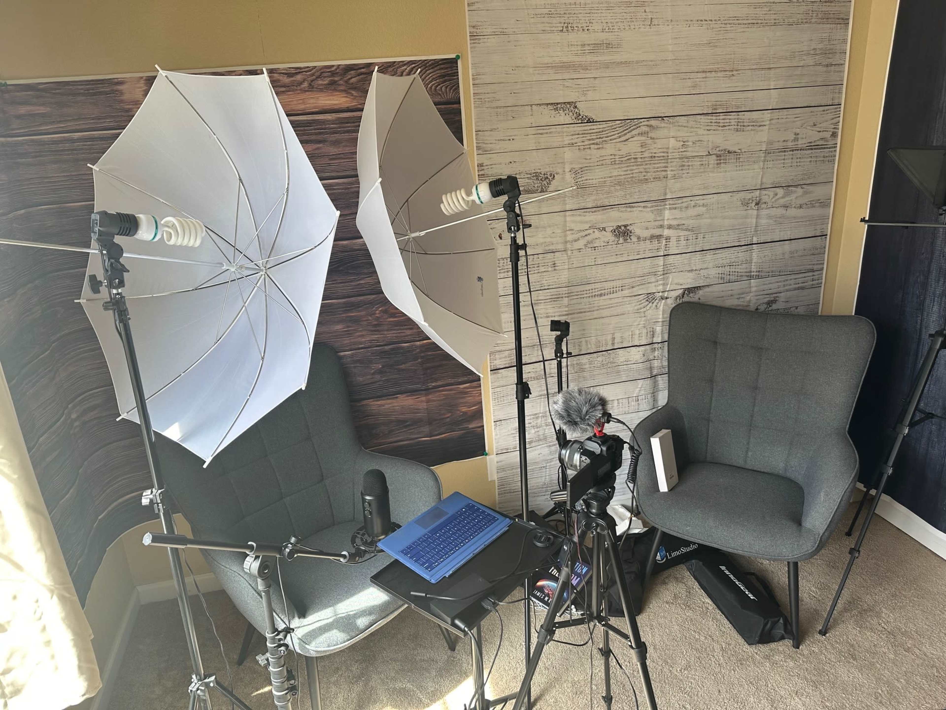 A setup for video recording features two umbrella lights, a laptop, and two chairs positioned in front of a wooden backdrop.