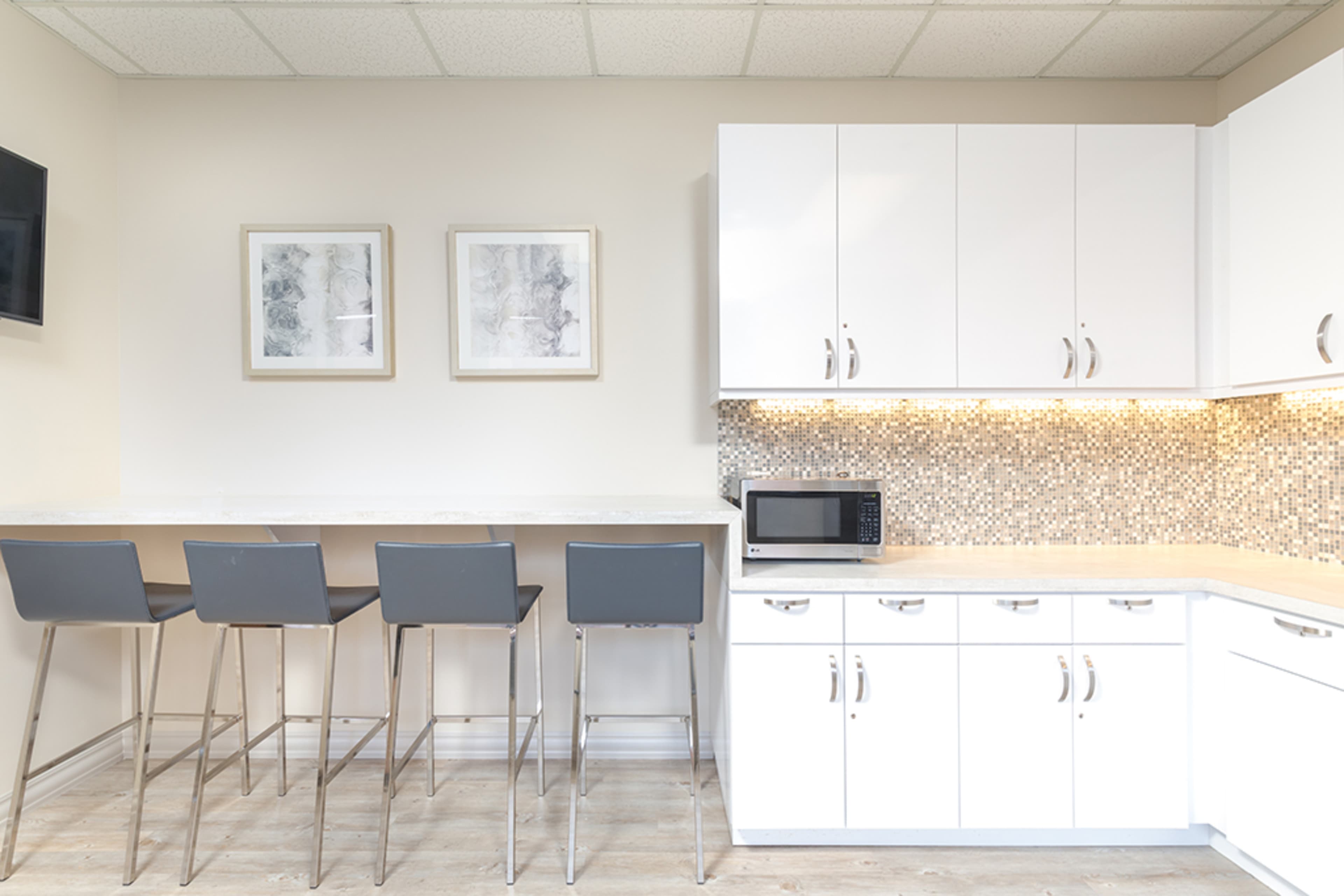 A modern kitchen features white cabinetry, a microwave, and a seating area with four gray bar stools against a light-colored wall.