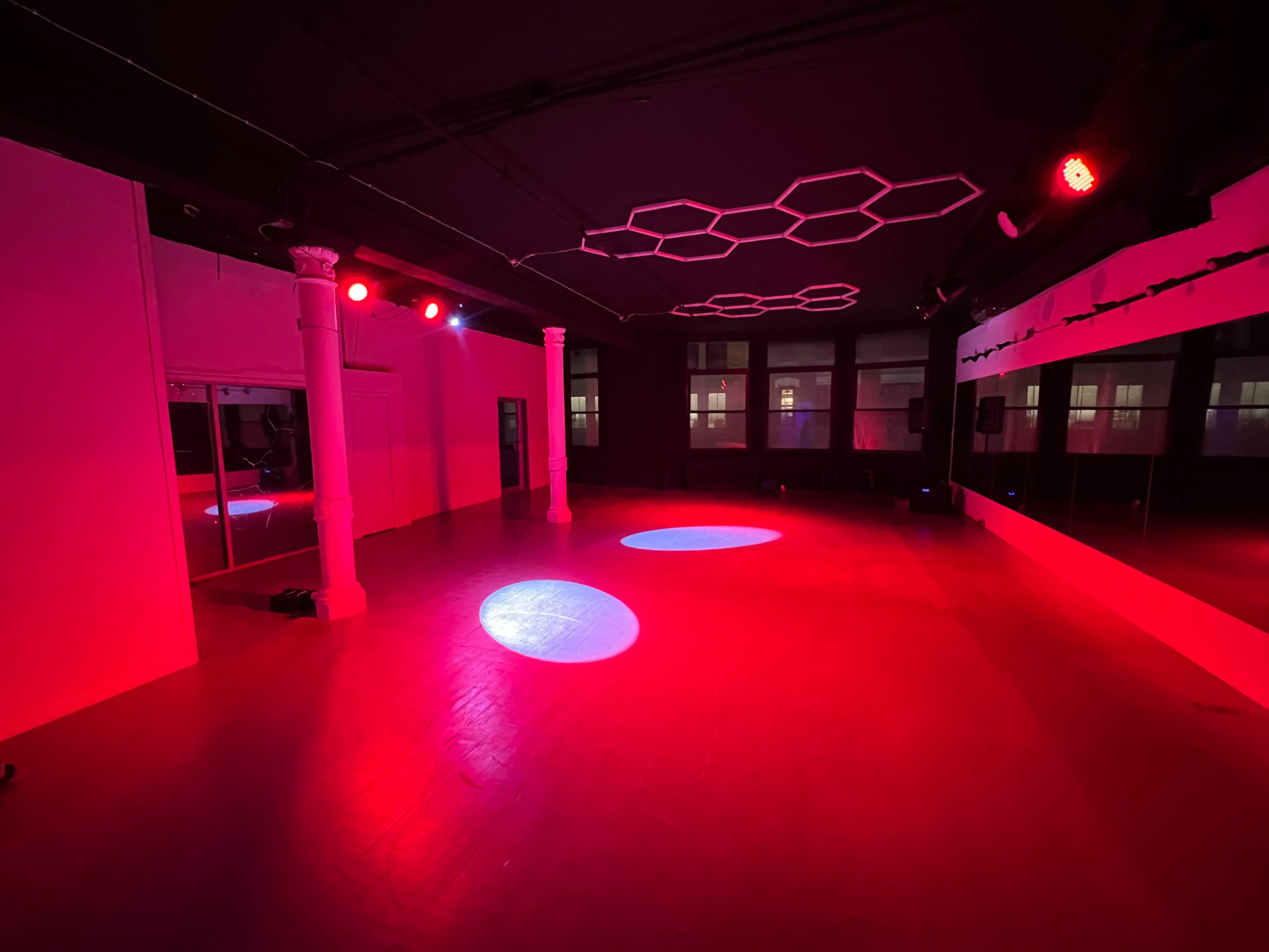 The image shows an empty dance studio with red lighting, featuring two circular light spots on the floor and mirrors along one wall.