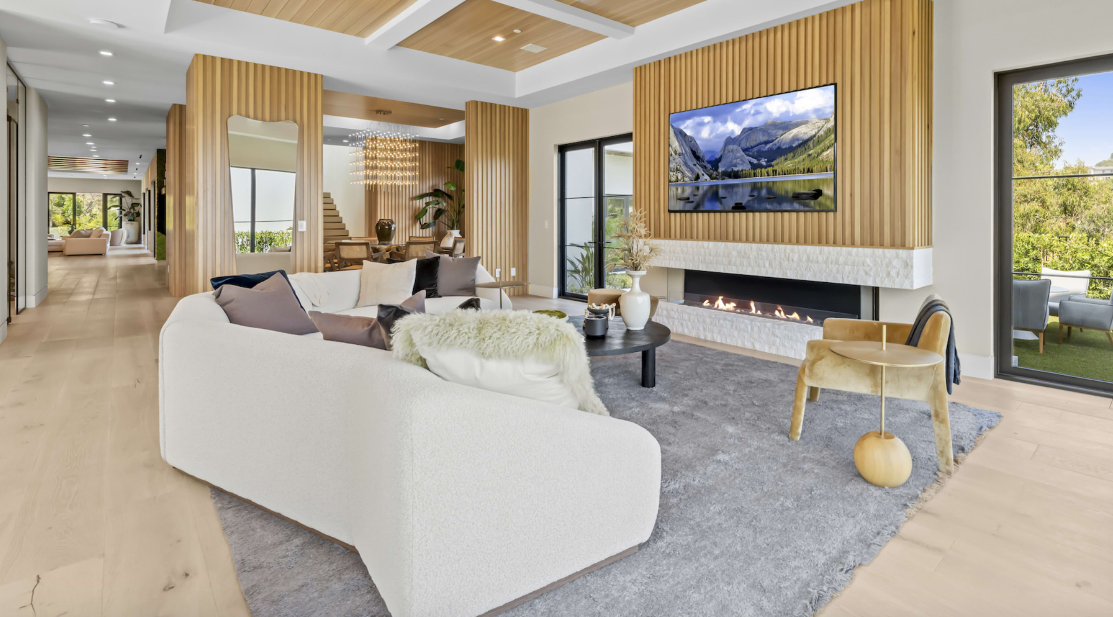 The image shows a modern living room with a white sofa, a round coffee table, and a wall-mounted television above a fireplace, all surrounded by wooden paneling and large glass doors leading to an outdoor space.