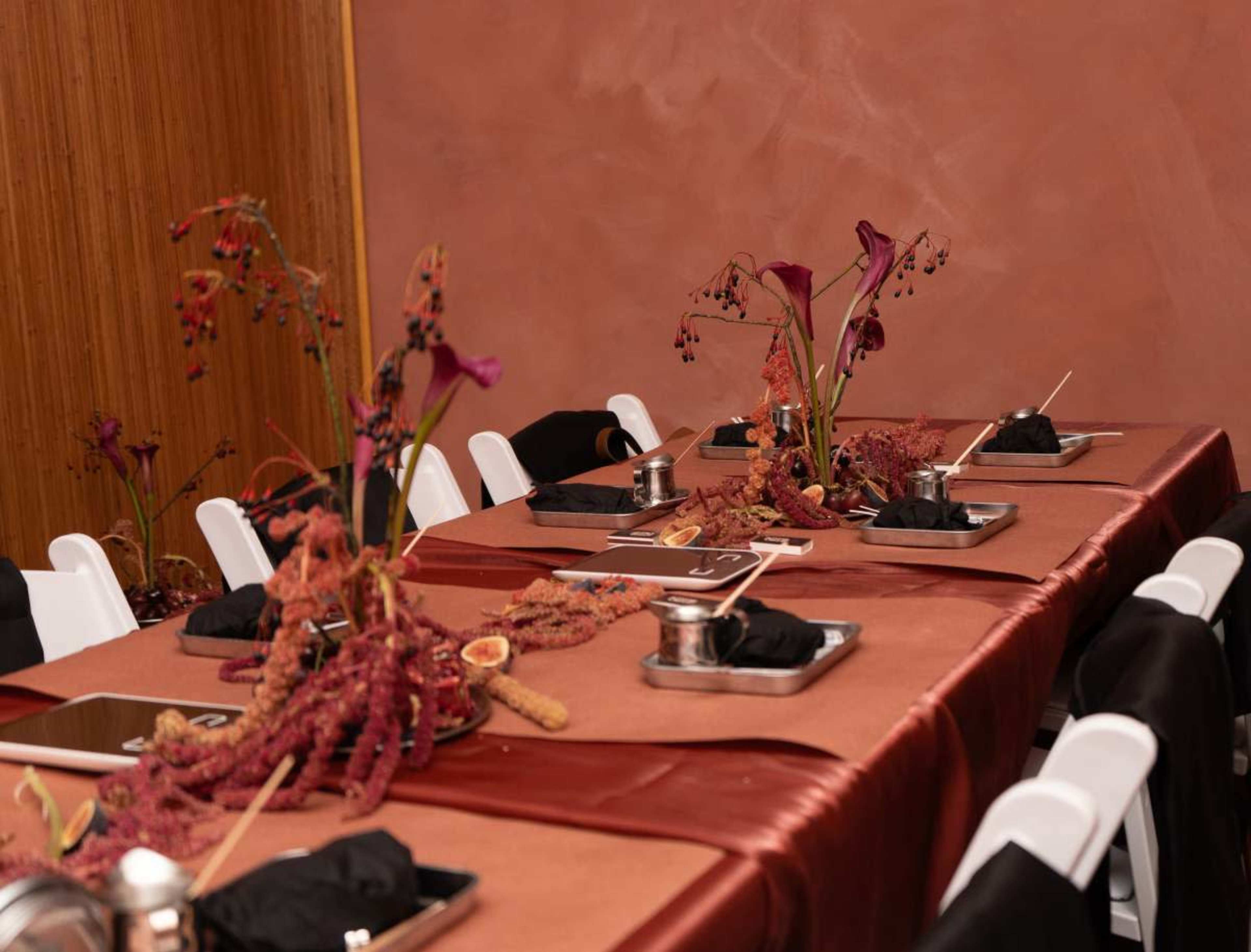 A long table is set for a gathering, featuring dark cloths, decorative arrangements of dried flowers, and metallic trays.