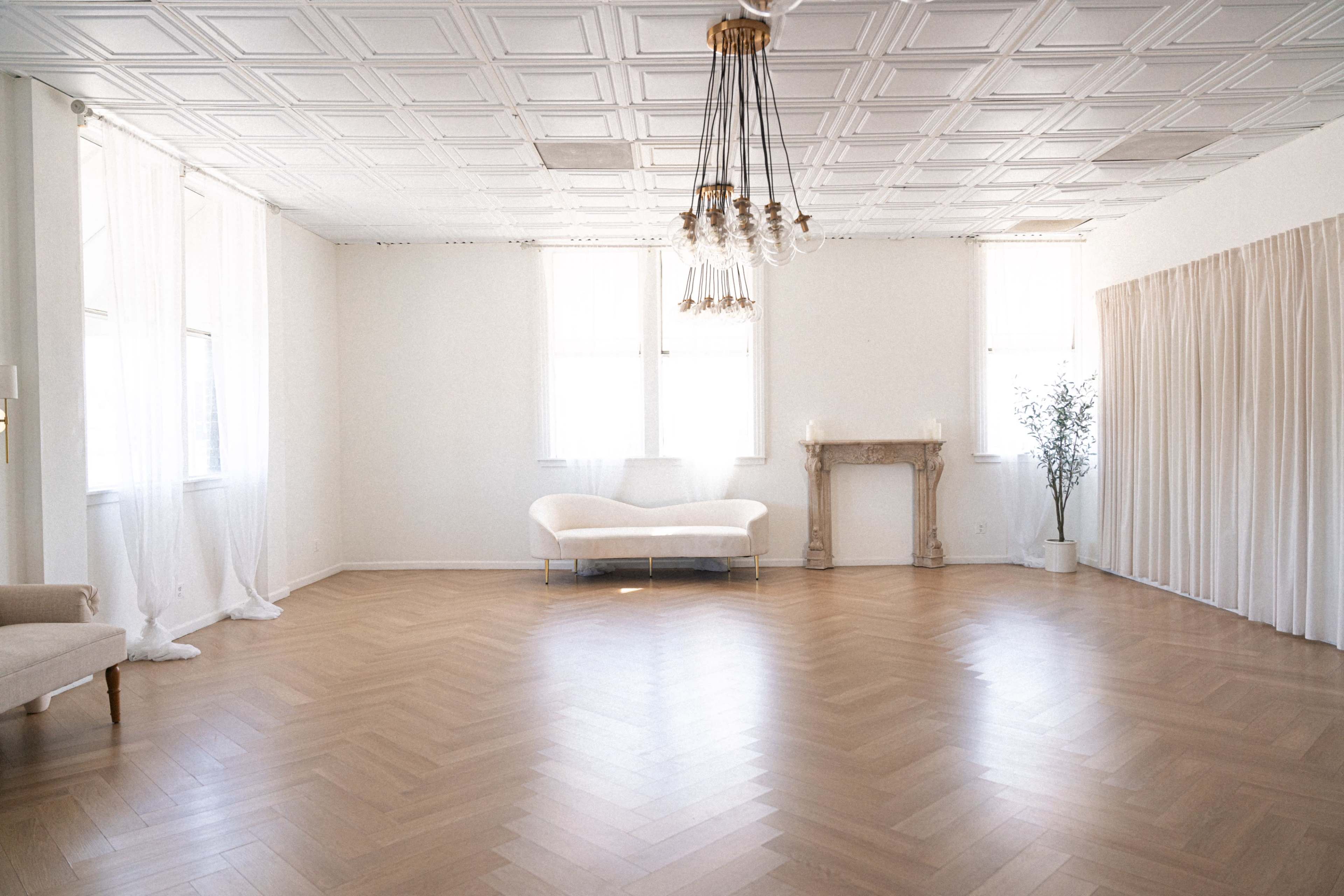 The image shows a spacious, well-lit room with light hardwood flooring, a chandelier hanging from the ceiling, a white sofa, and a decorative fireplace.