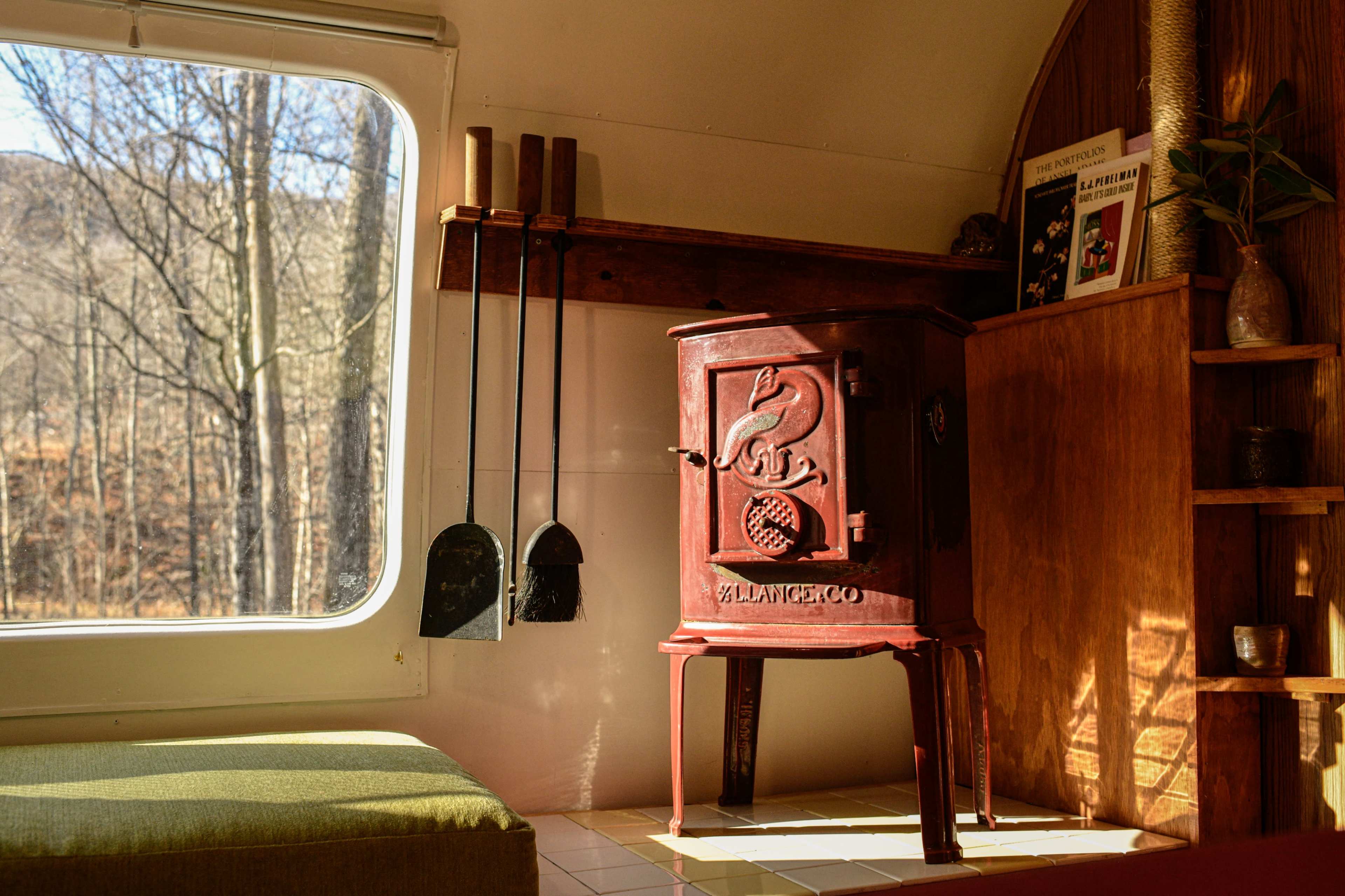 A vintage red wood stove positioned next to a green cushion and a window with a view of bare trees outside.