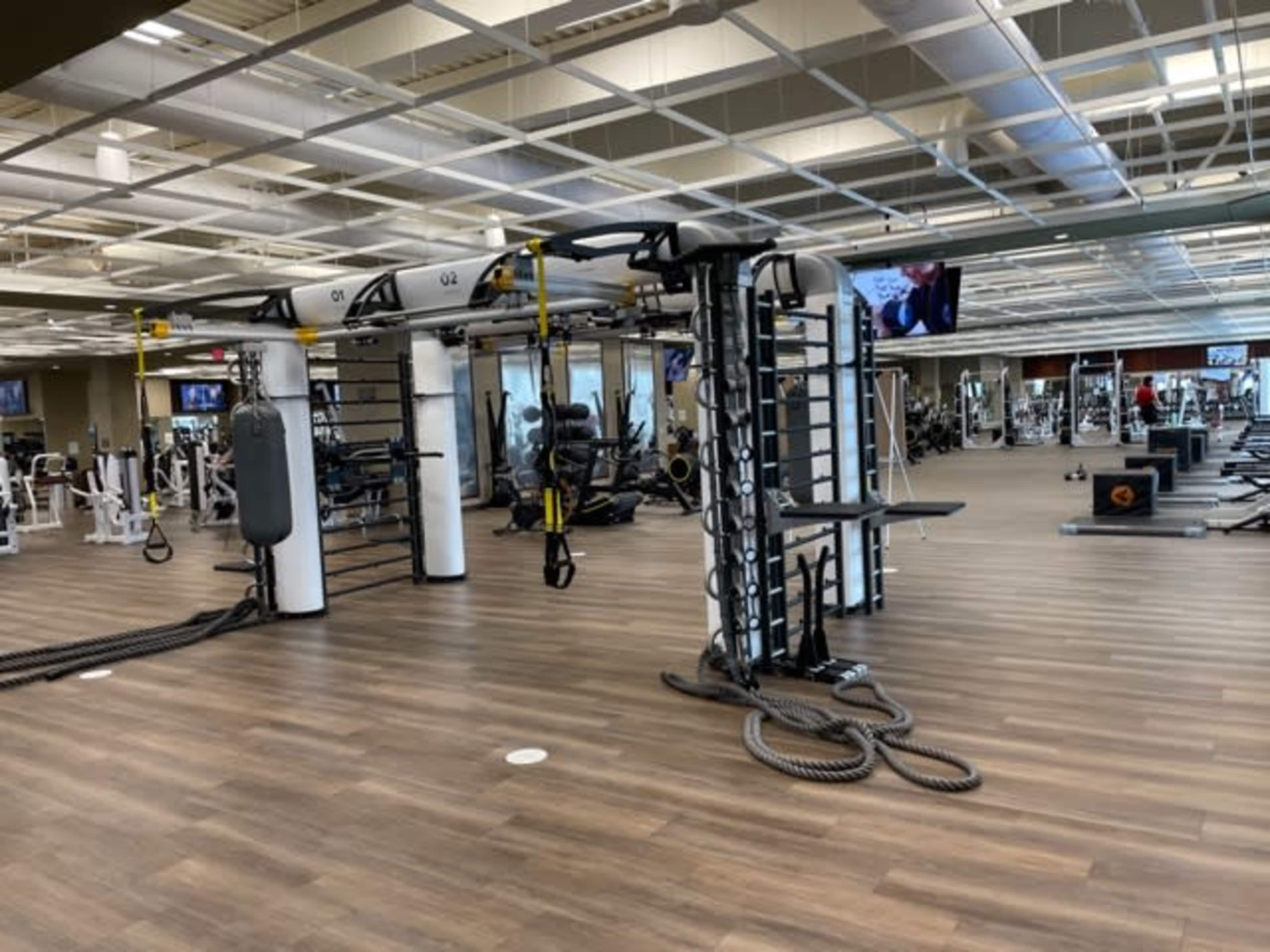 The image shows a spacious gym interior with various exercise equipment, including cable machines and free weights, set against a backdrop of wooden flooring and ample lighting.