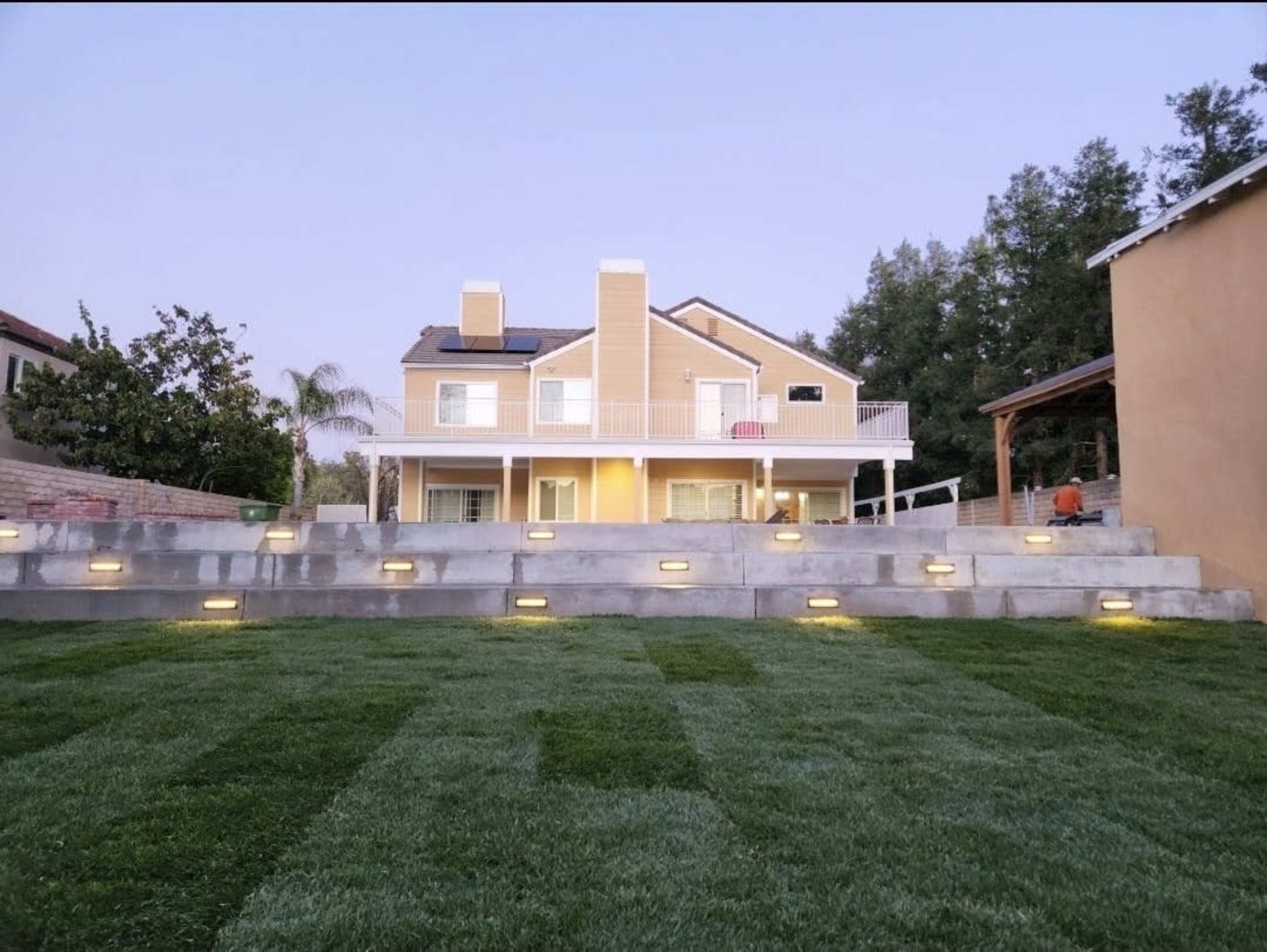 The image shows a two-story yellow house with a balcony and a landscaped garden featuring tiered lawn and lighting.