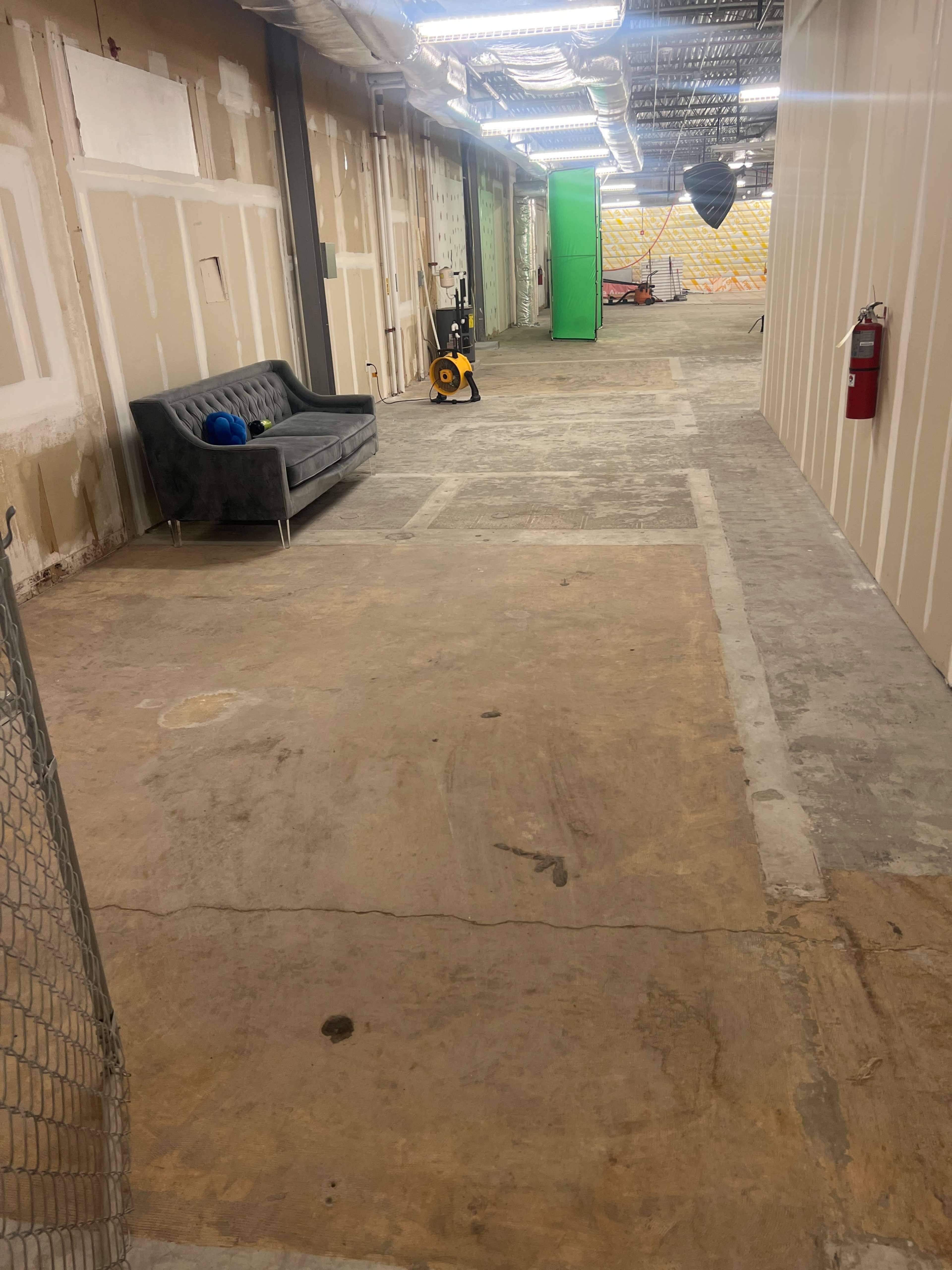 The image shows a long, empty hallway with bare walls, an unadorned concrete floor, and a couch on one side.