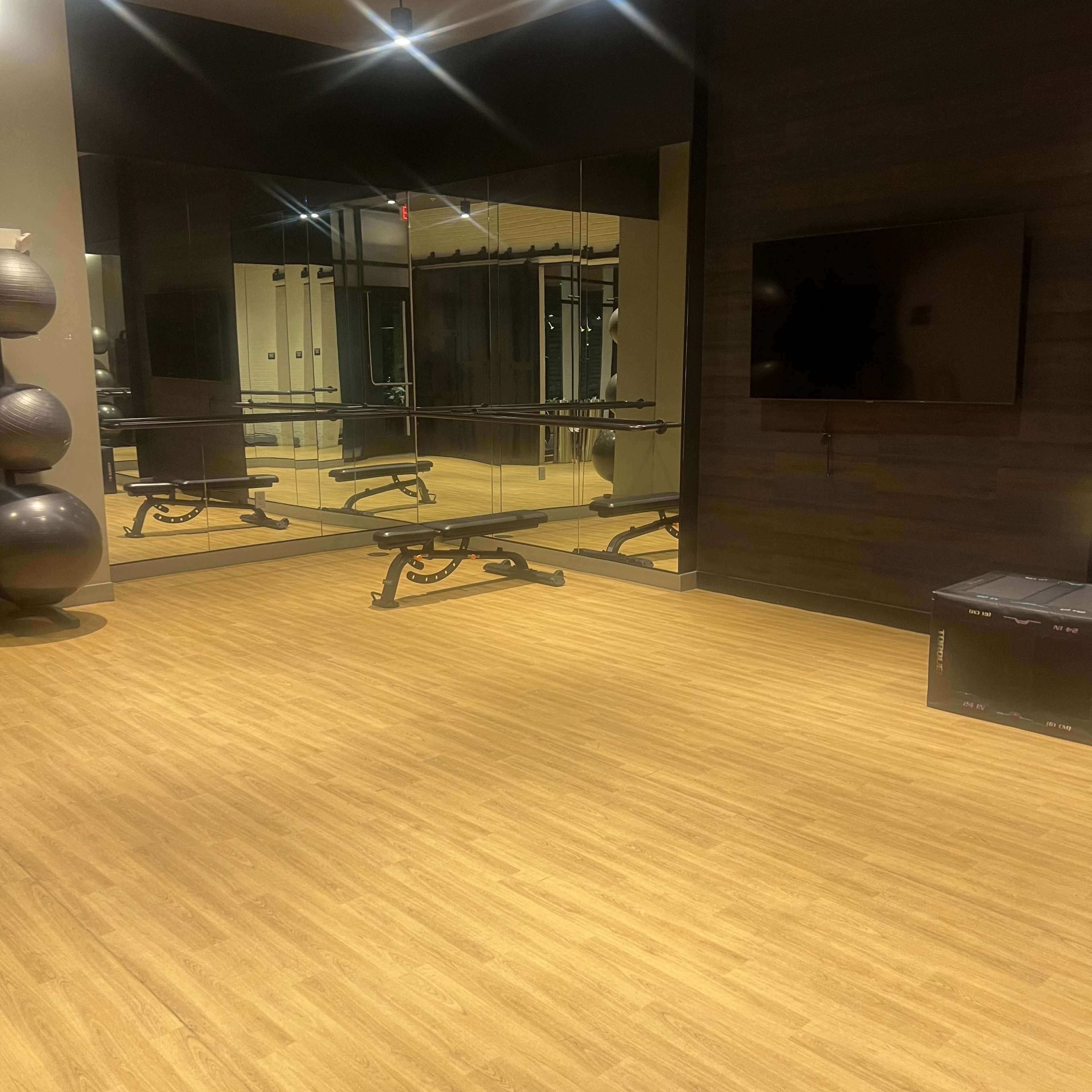 The image shows a modern gym interior with a reflective wall of mirrors, a weight bench, and exercise equipment, all set on a wooden floor.