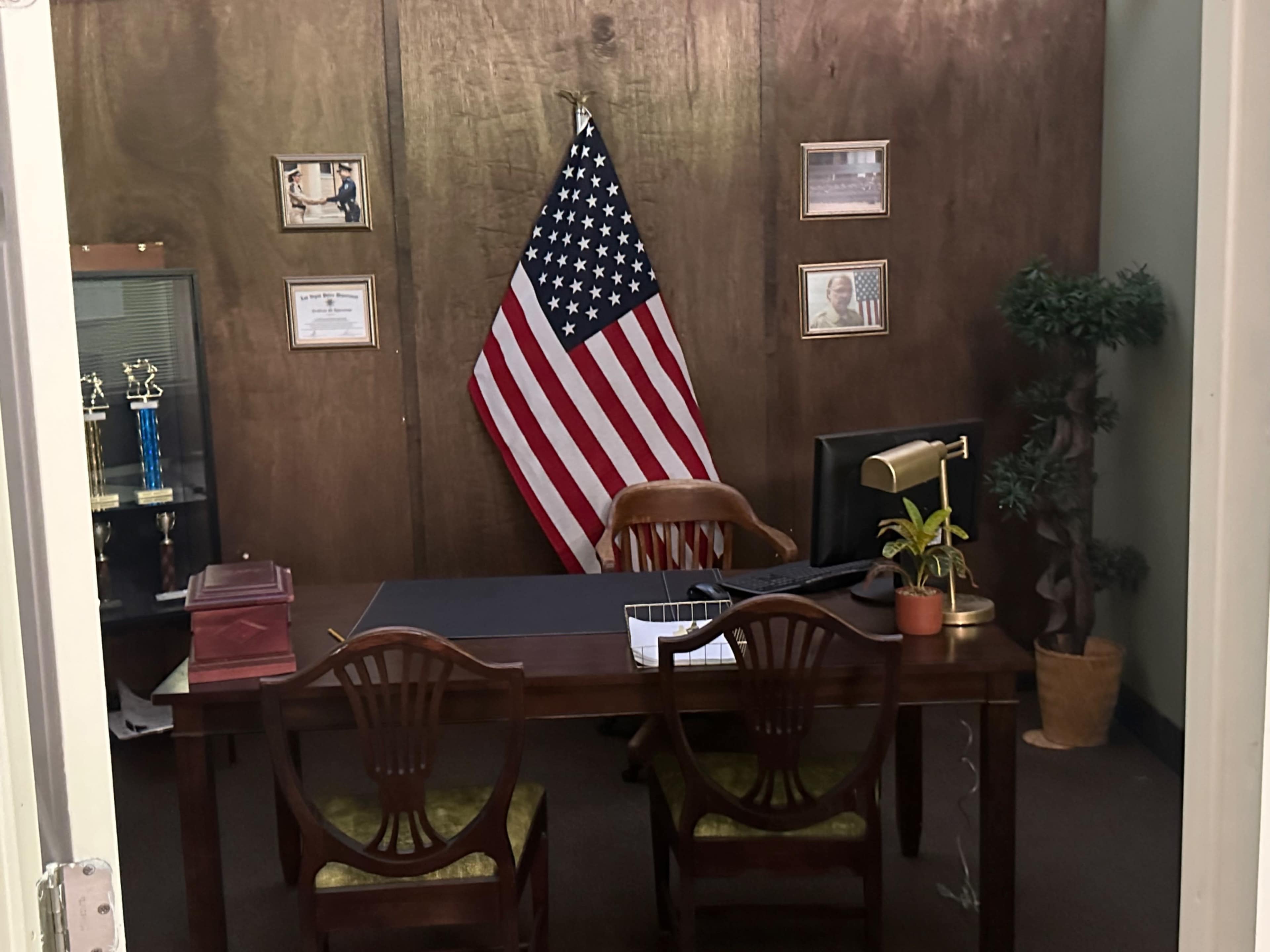 The image depicts an office setup featuring a wooden desk, two chairs, an American flag, a potted plant, and various framed certificates on the wall.