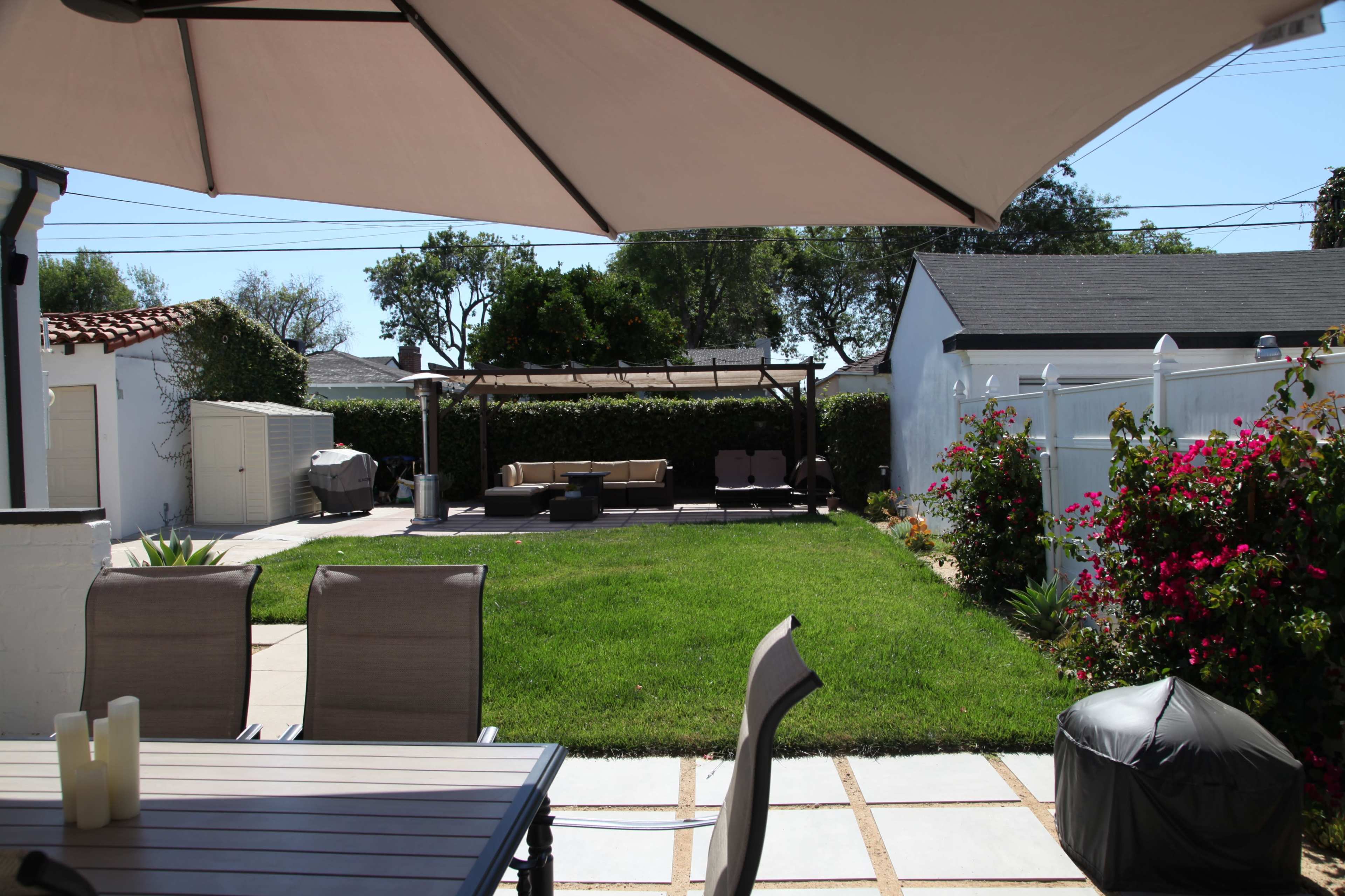 A patio area features a table and chairs in the foreground, with a grassy yard and seating area in the background under a large shade umbrella.