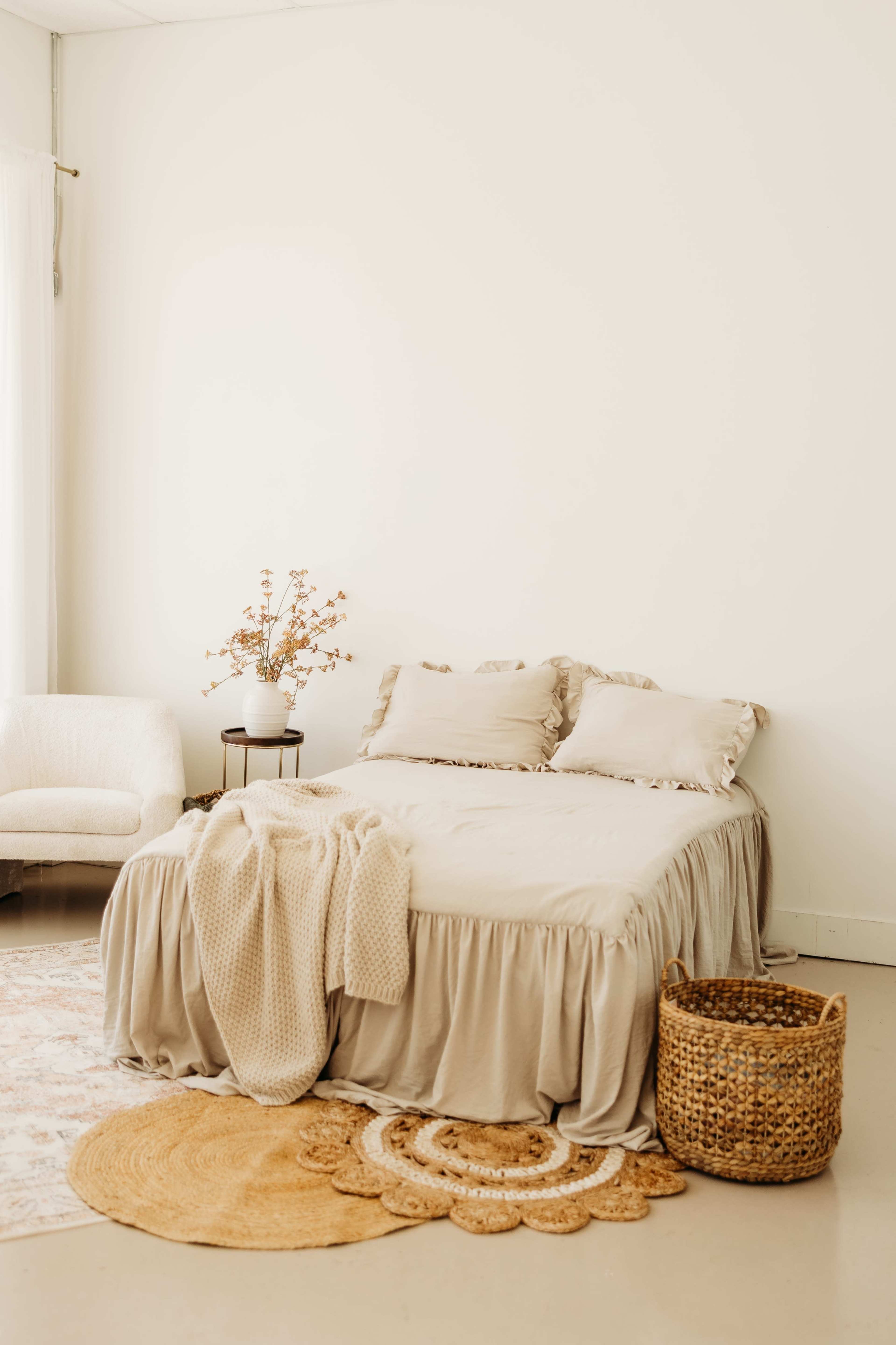 The image shows a softly decorated bedroom featuring a bed with a light beige duvet, two pillows, a woven basket, and a small chair beside a table with a simple floral arrangement.