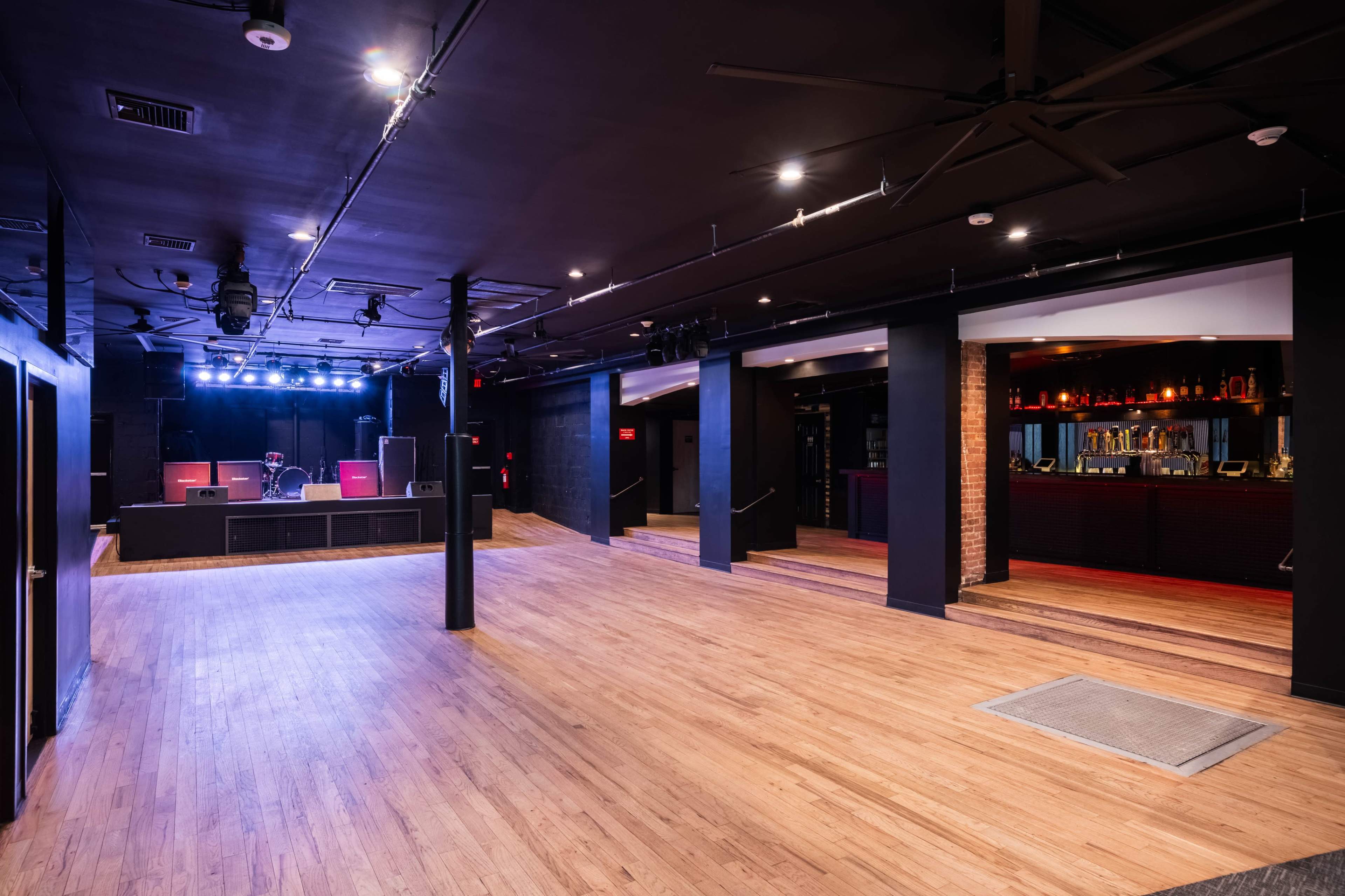 The image shows an empty music venue with a stage at the front, wooden floor, and a bar area to the right.