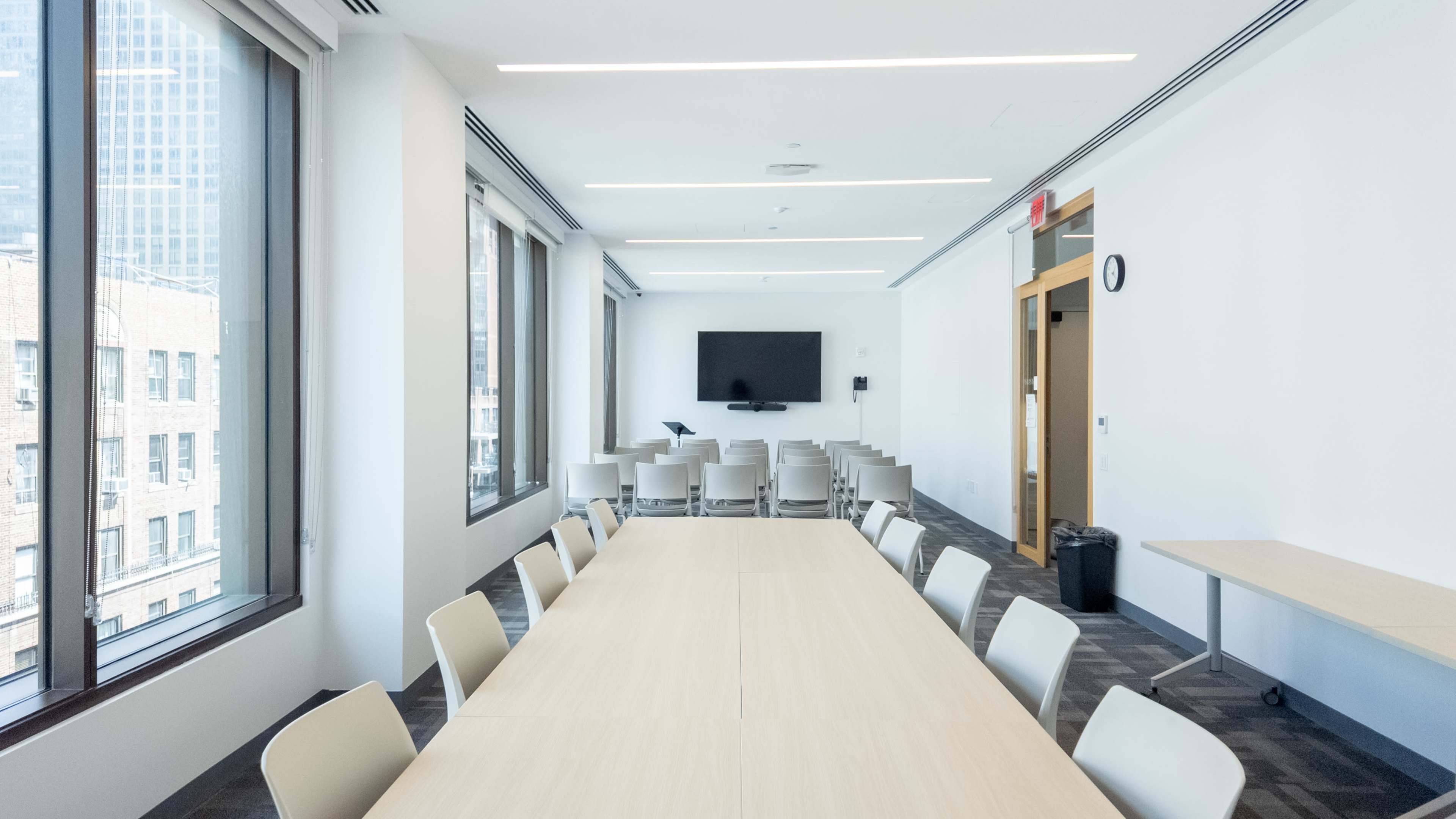 The image shows a modern conference room featuring a long table, several chairs, large windows, and a wall-mounted television.