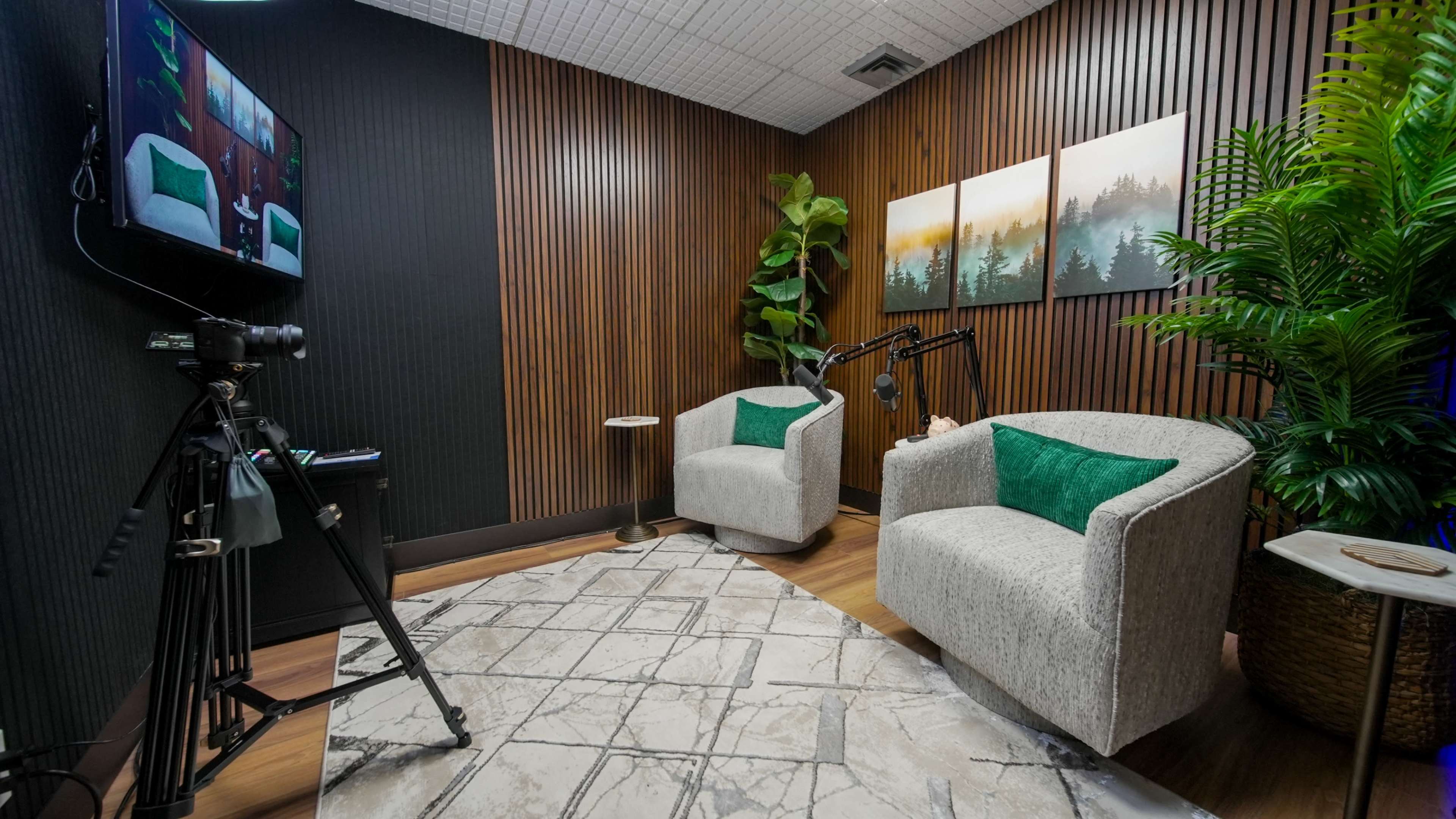 The image shows a small recording studio featuring two chairs with green accents, a camera on a tripod, and a wall adorned with landscape artwork.