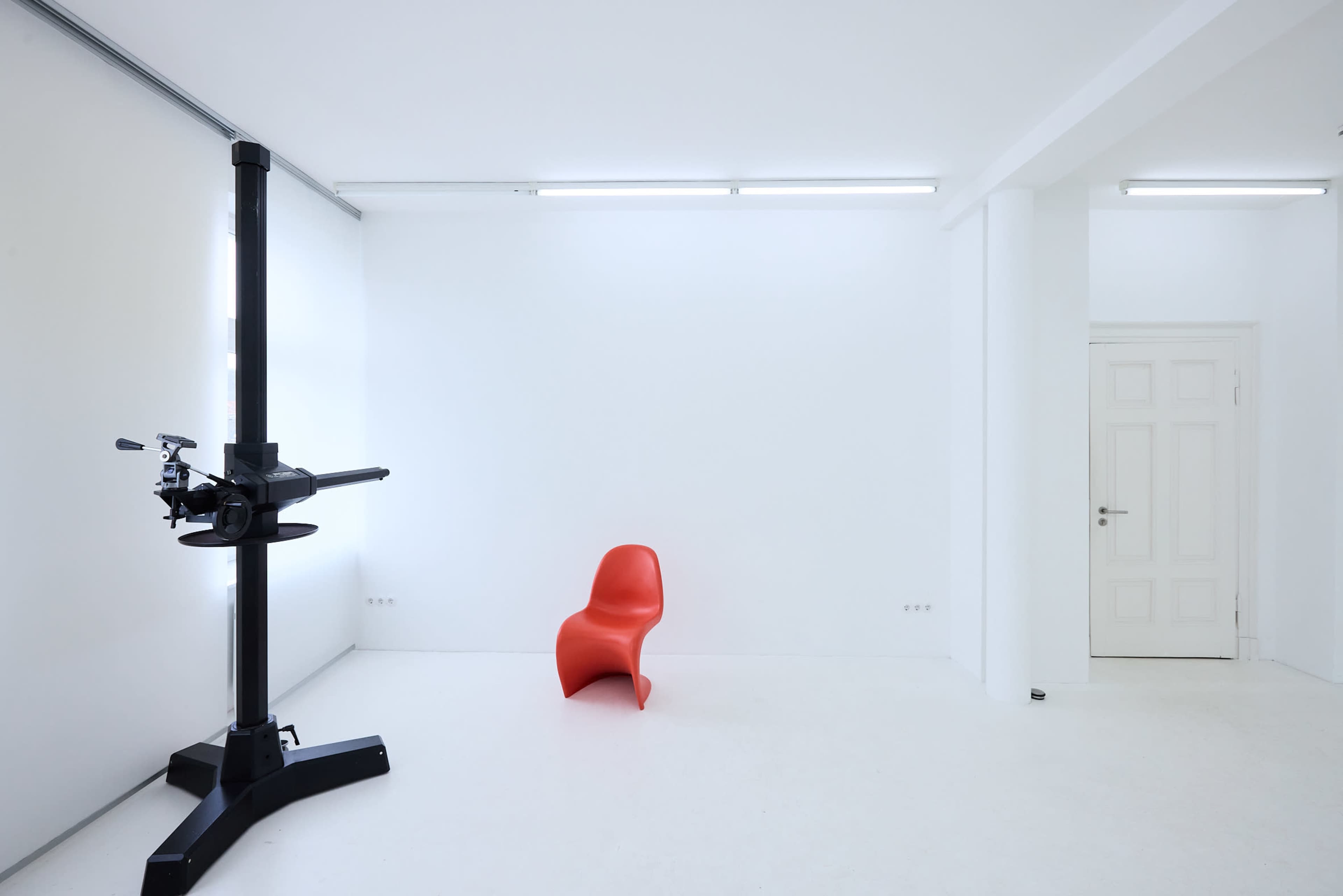The image shows a minimalist white room with a black machine on the left and a red chair on the right.