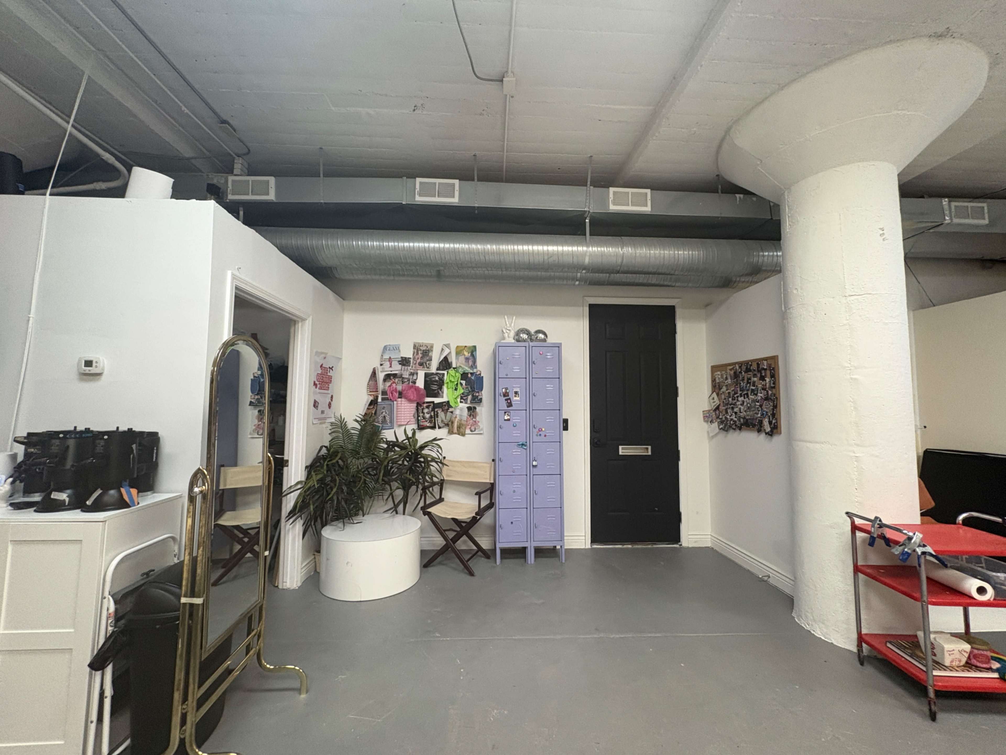 The image shows a simple, industrial-style room featuring a tall white column, a purple locker, and a mirrored stand along with some wall art and furniture.