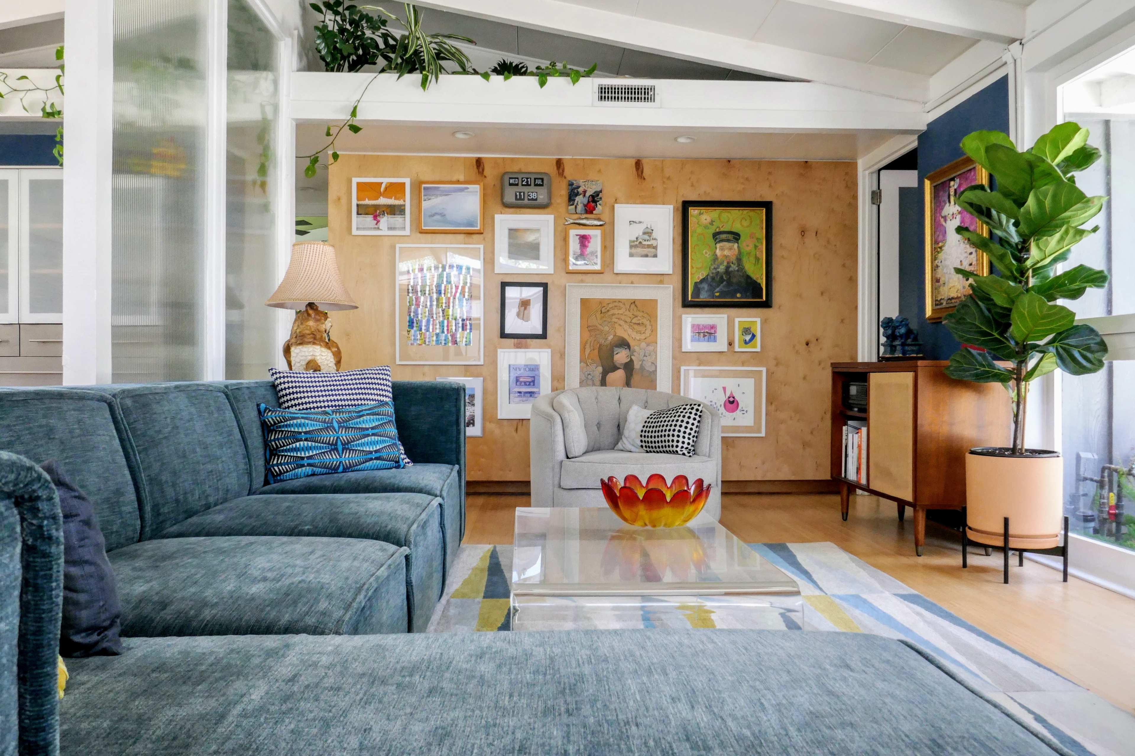 The image shows a modern living room with a sectional sofa, a coffee table, a variety of framed artwork on the walls, and a potted plant near a large window.