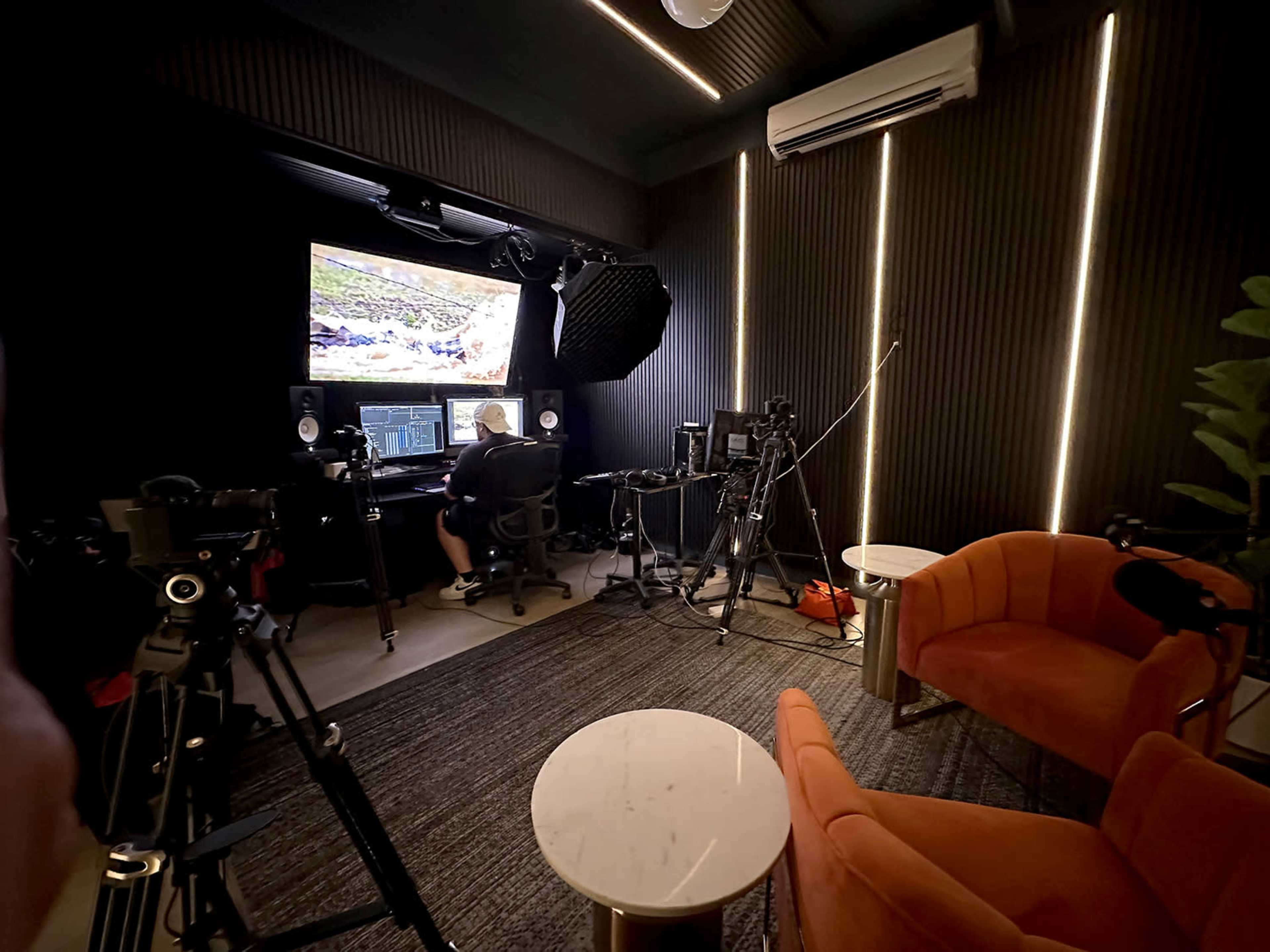The image shows a recording studio equipped with audio and video equipment, featuring a desk with a computer display, and two orange chairs placed nearby.