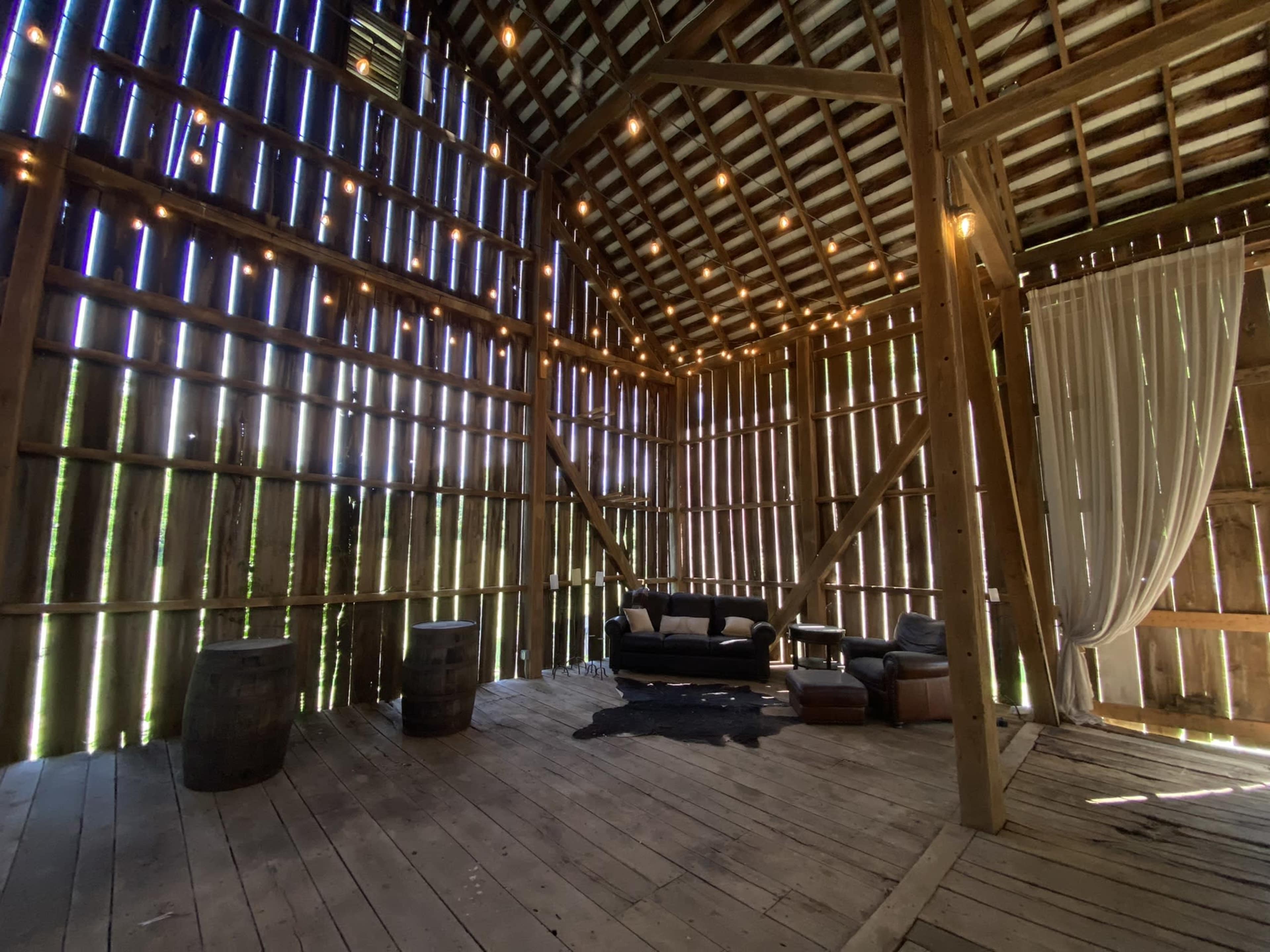 The interior of a rustic barn features spaced wooden slats, string lights, and a seating area with a sofa and leather chair on a wooden floor.
