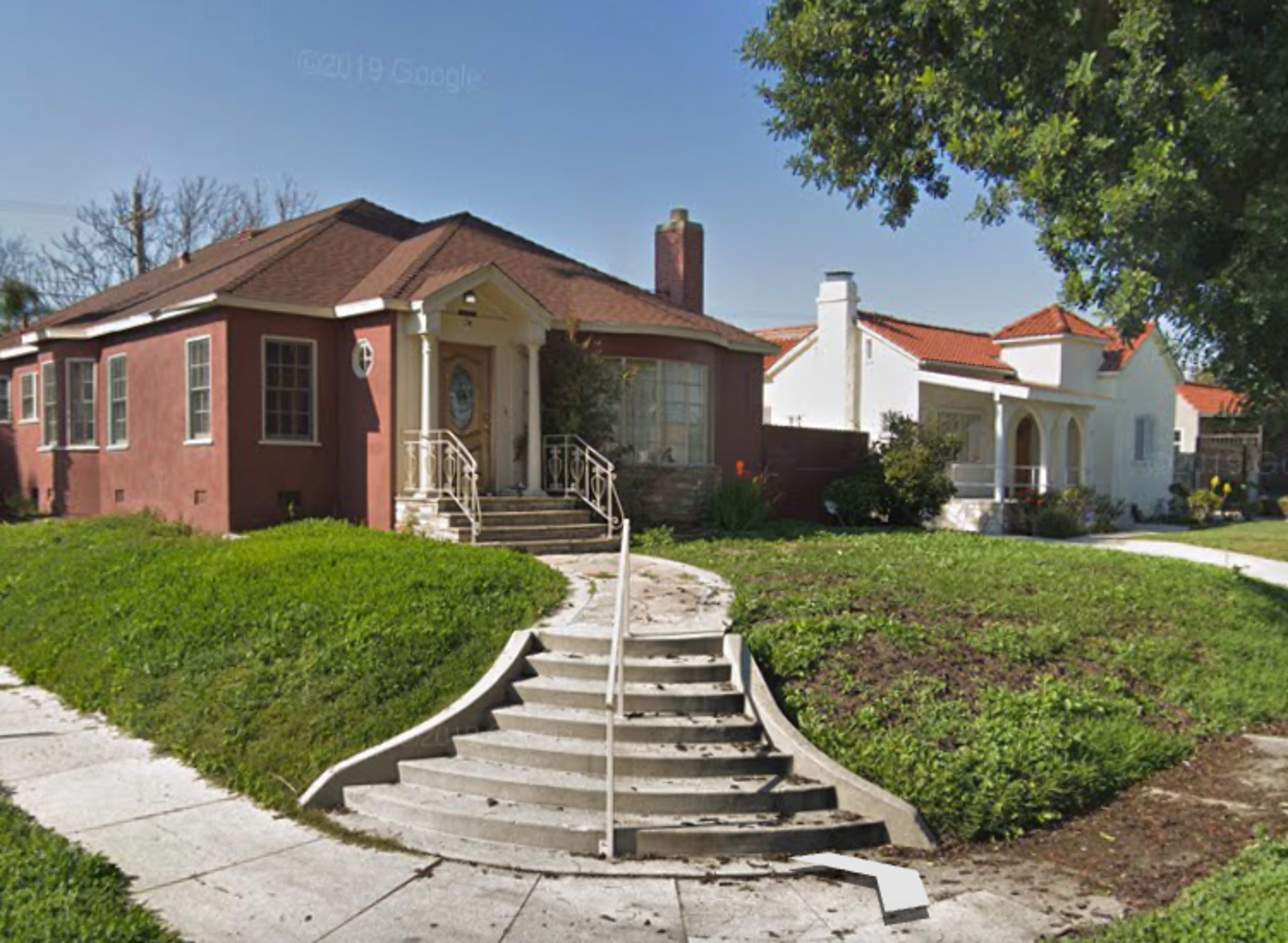 A single-story house with a red exterior and a prominent staircase leading to the front entrance, situated next to a white house with a similar structure on a grassy corner lot.