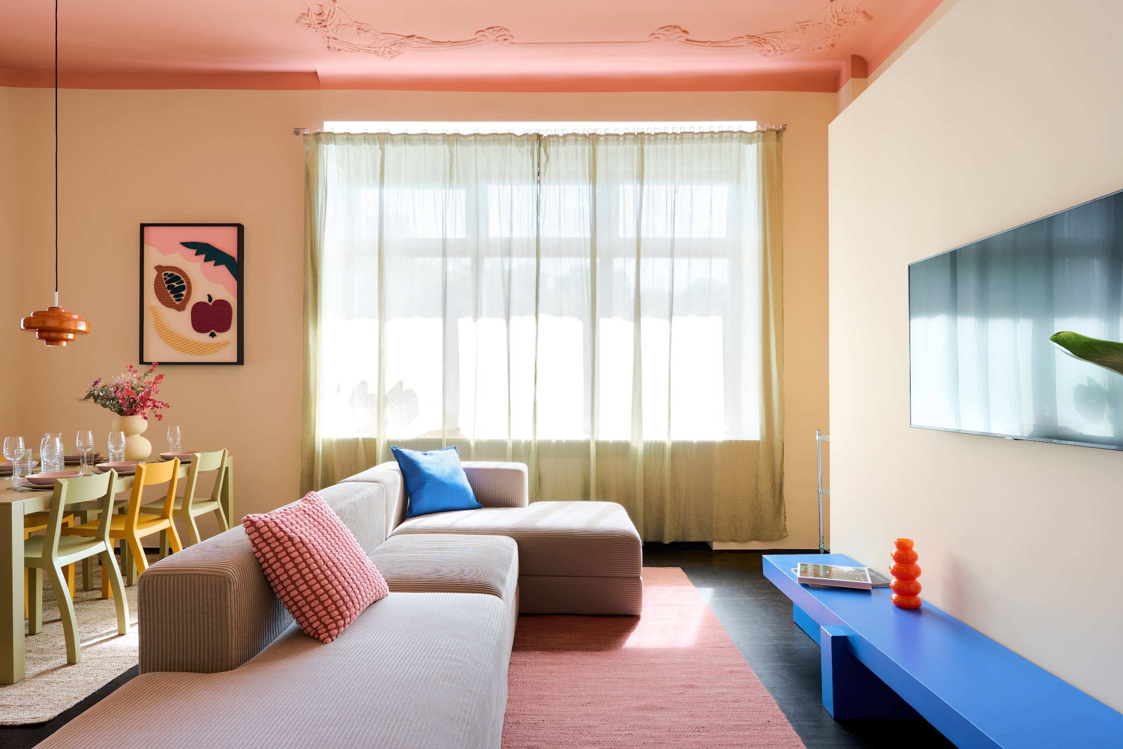 The image shows a modern living room with a beige sofa, a blue TV stand, and a dining area featuring a table with green chairs and a vase of flowers, illuminated by natural light from large windows.