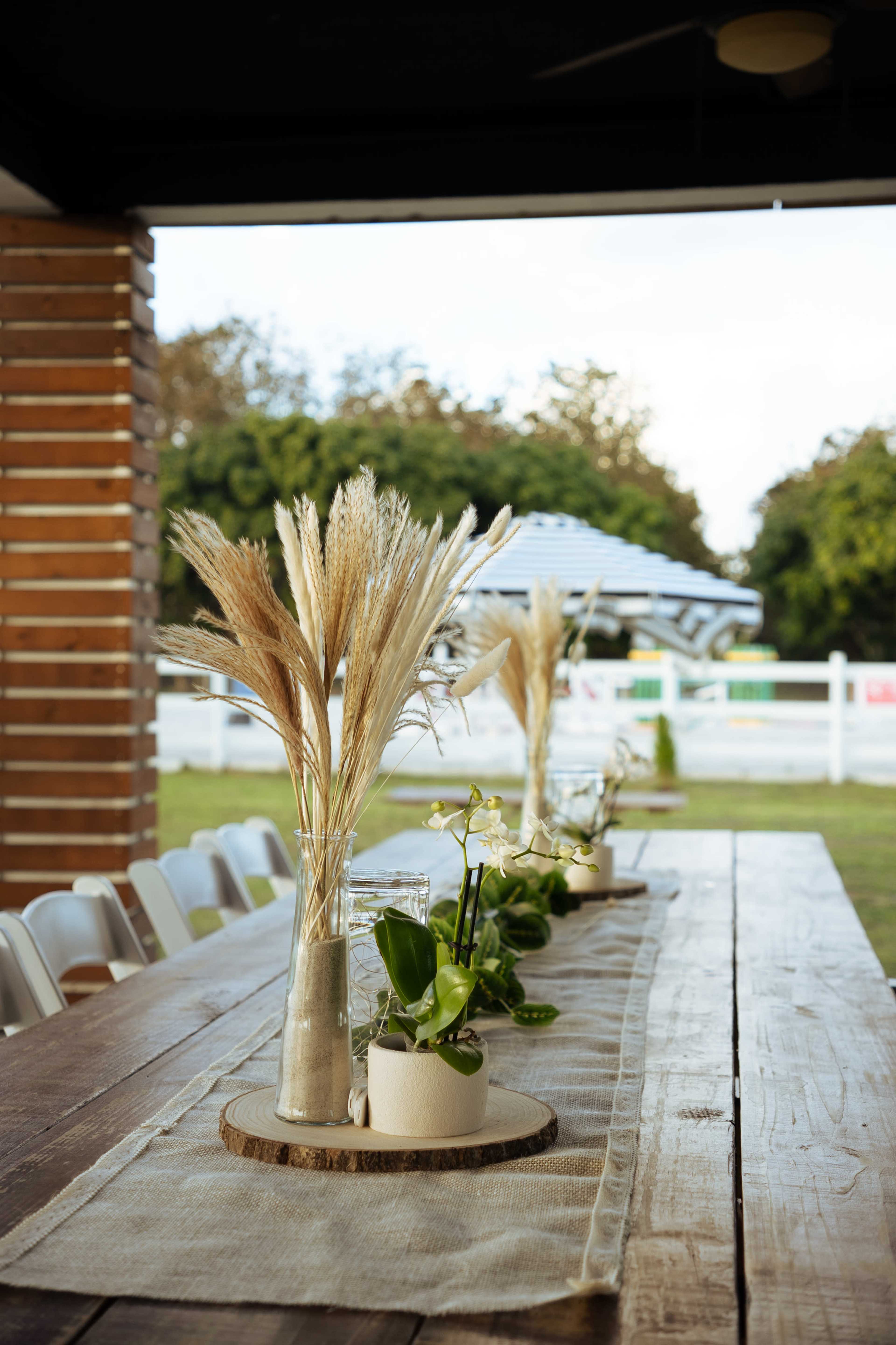 A long wooden table is decorated with vases holding dried grasses and small potted plants, set outdoors under a covered area.