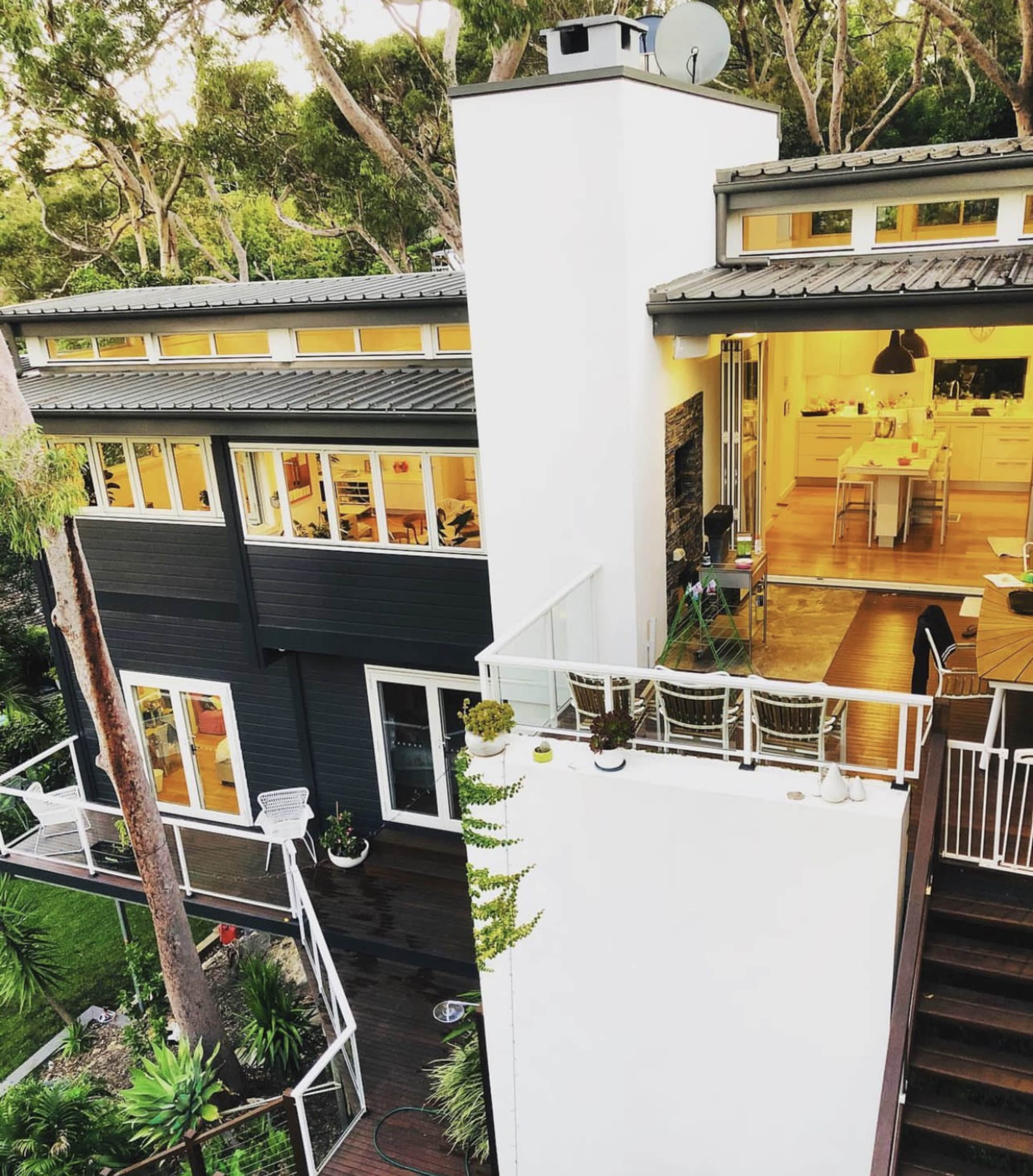 The image shows a modern two-story house with large windows surrounded by lush greenery, featuring a balcony and an outdoor seating area.