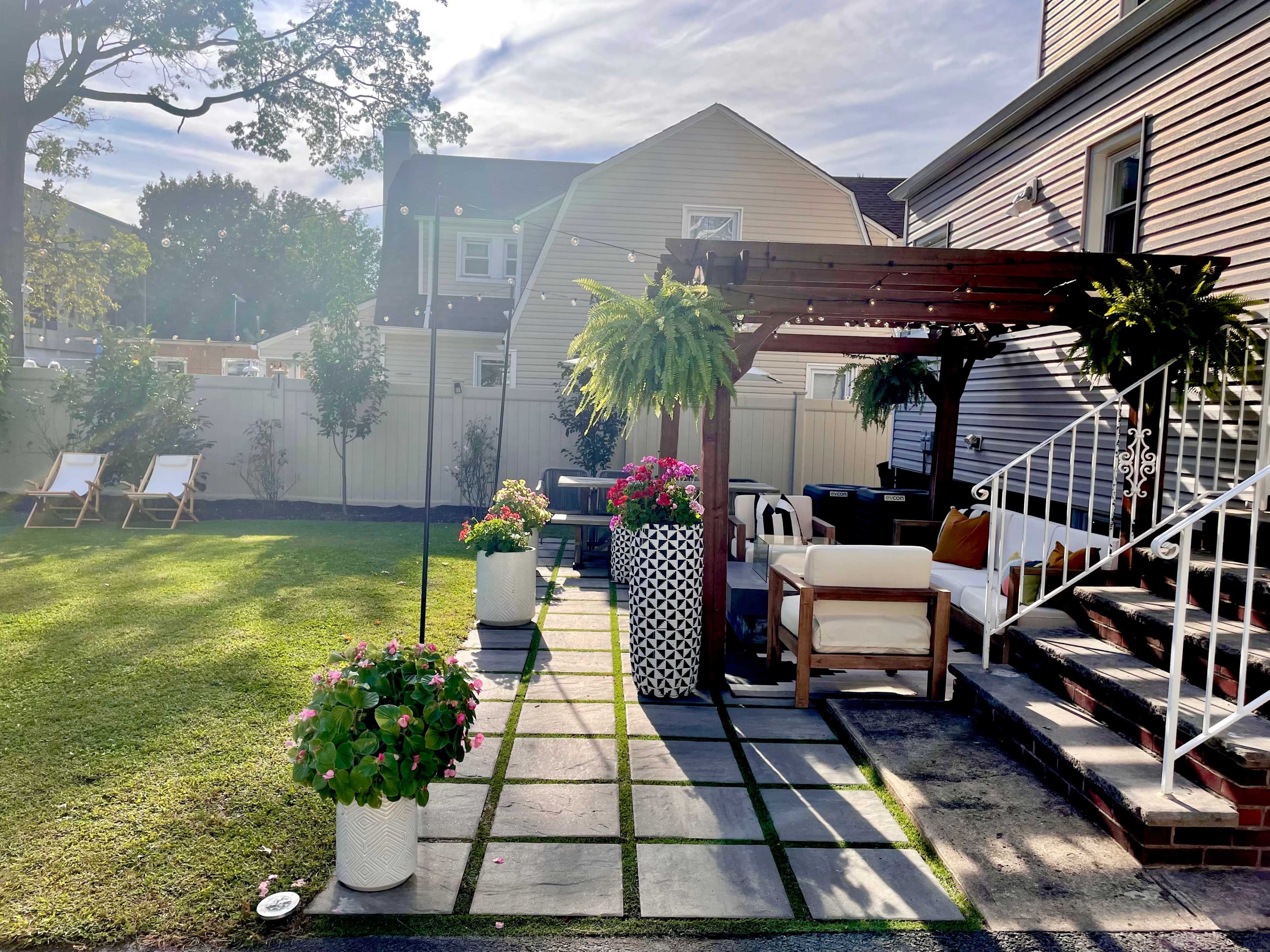 A landscaped backyard featuring a patio with a pergola, potted plants, and two lounge chairs.
