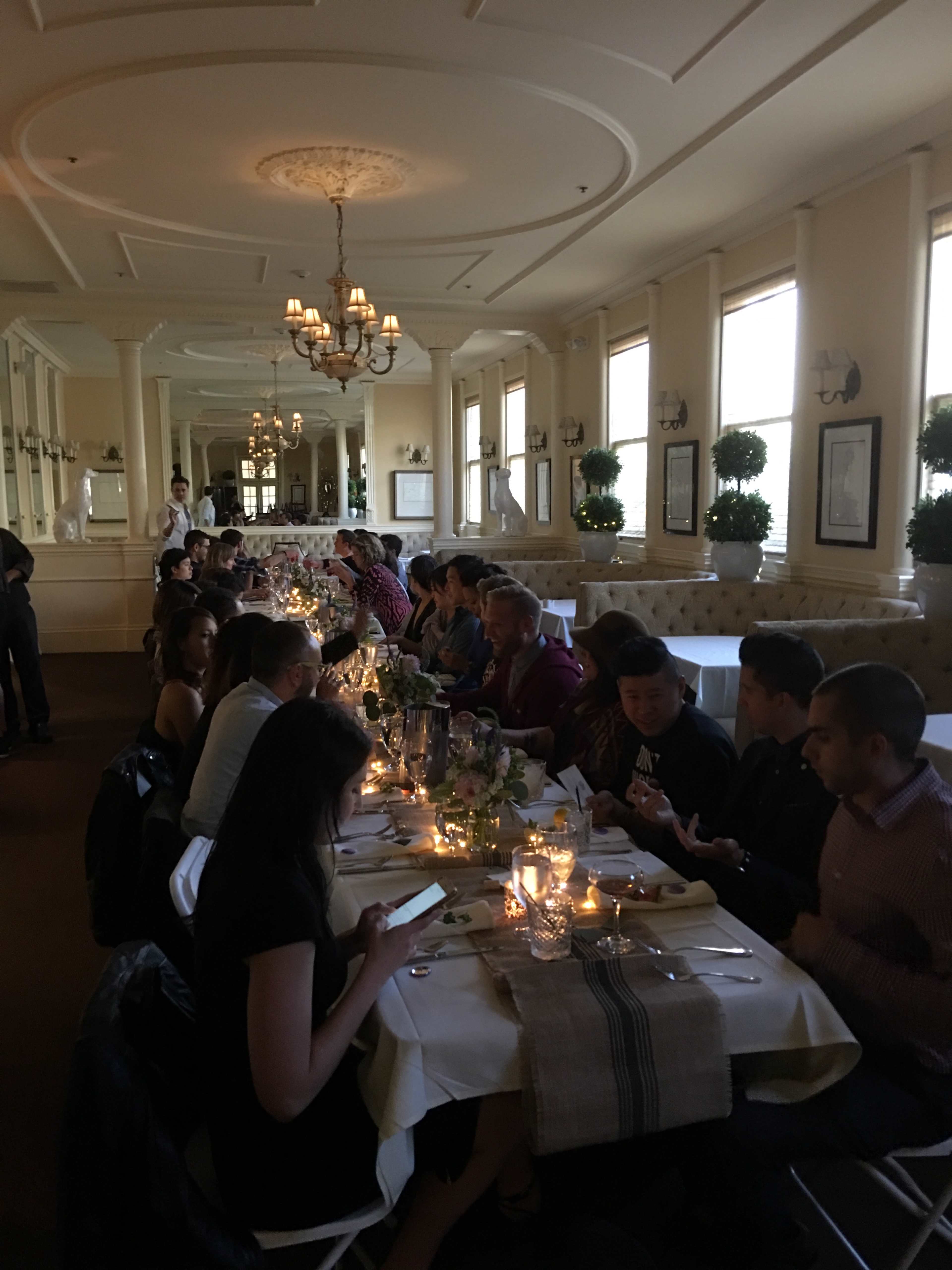 A long dining table is set for a gathering in a well-lit restaurant, with guests seated on both sides and candles illuminating the space.