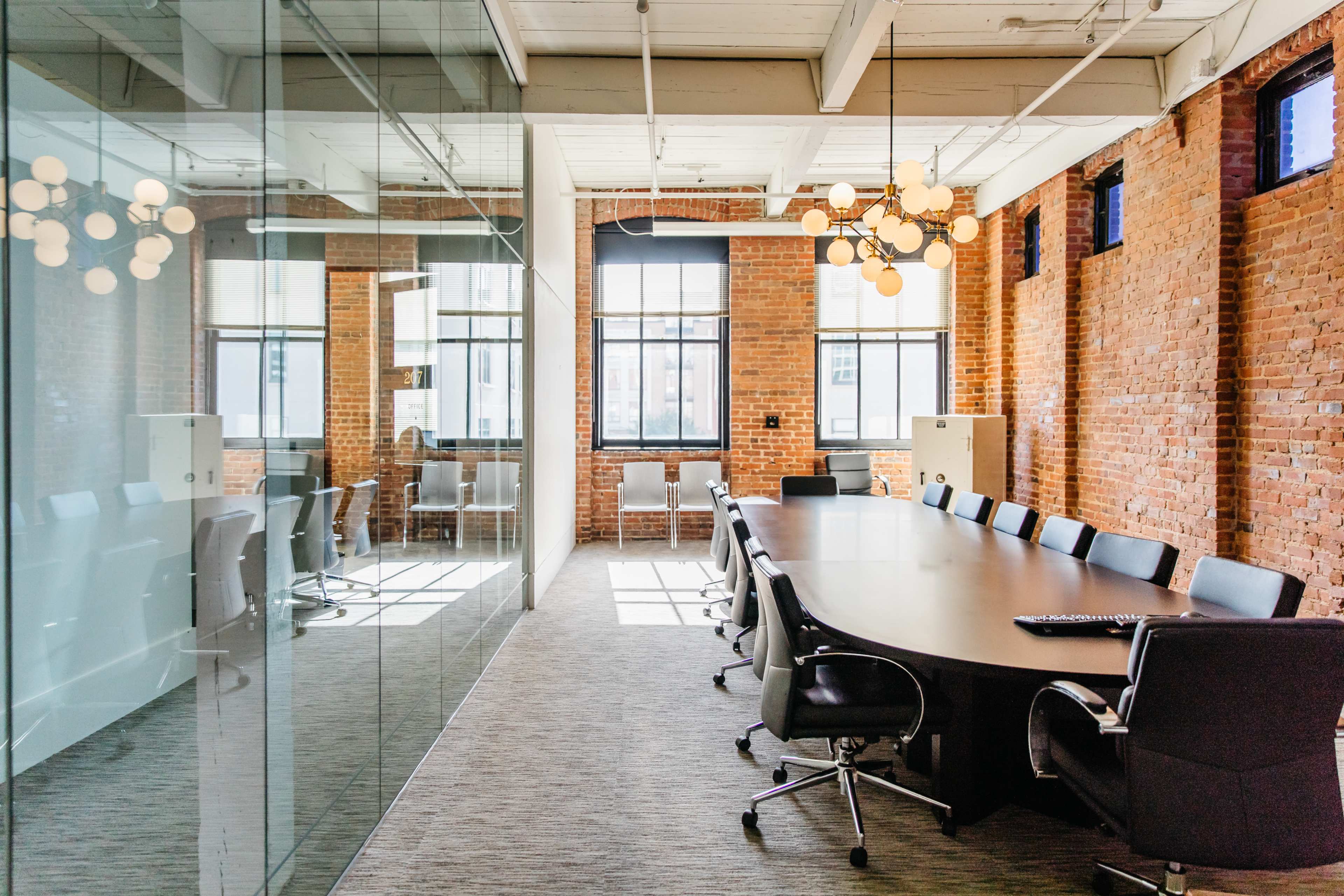 A conference room features a long table surrounded by black chairs, with a glass wall reflecting the space and brick walls in the background.