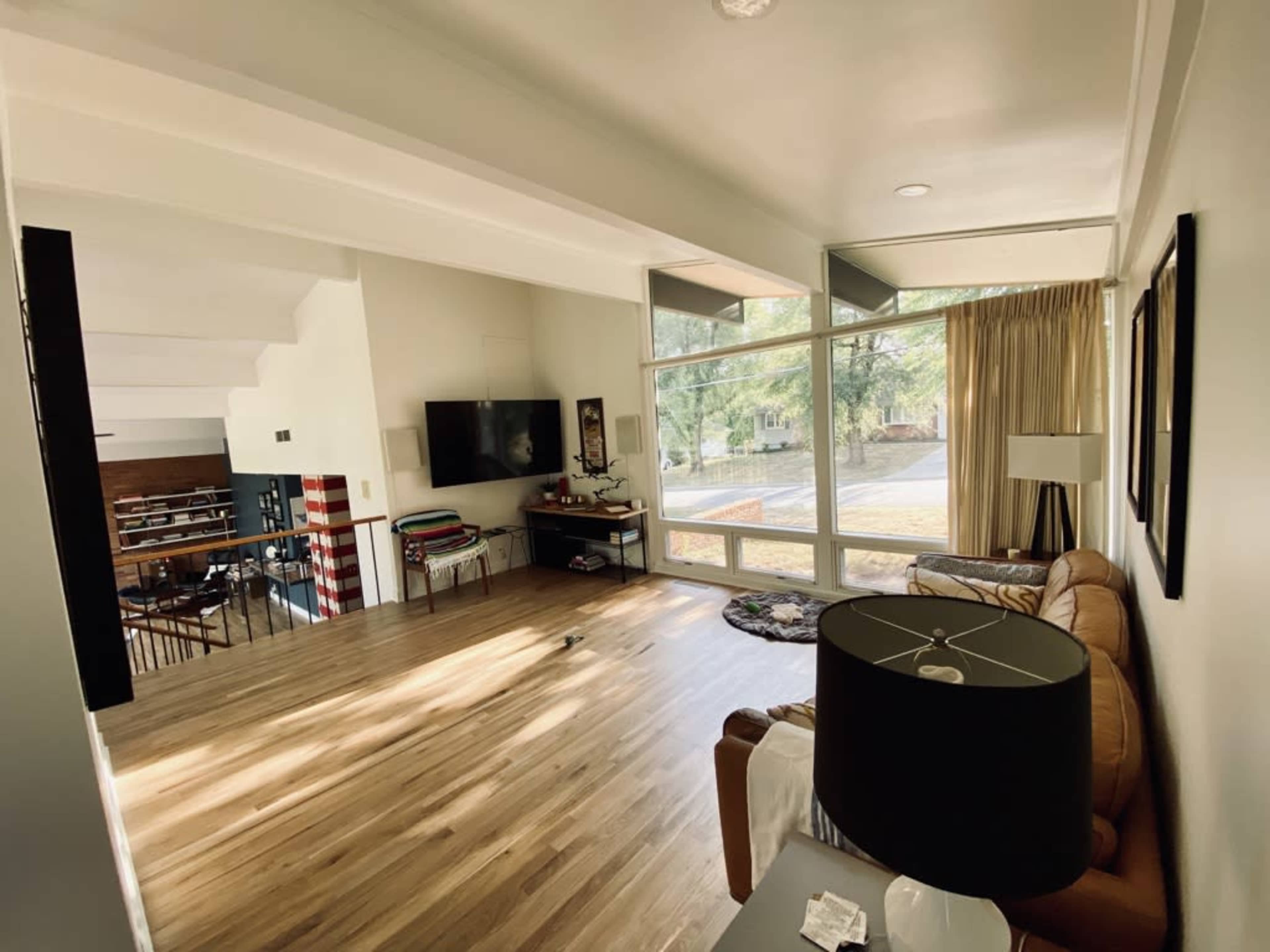 A spacious living room features a large window, hardwood floors, a sofa, a TV mounted on the wall, and a small lamp on a table.