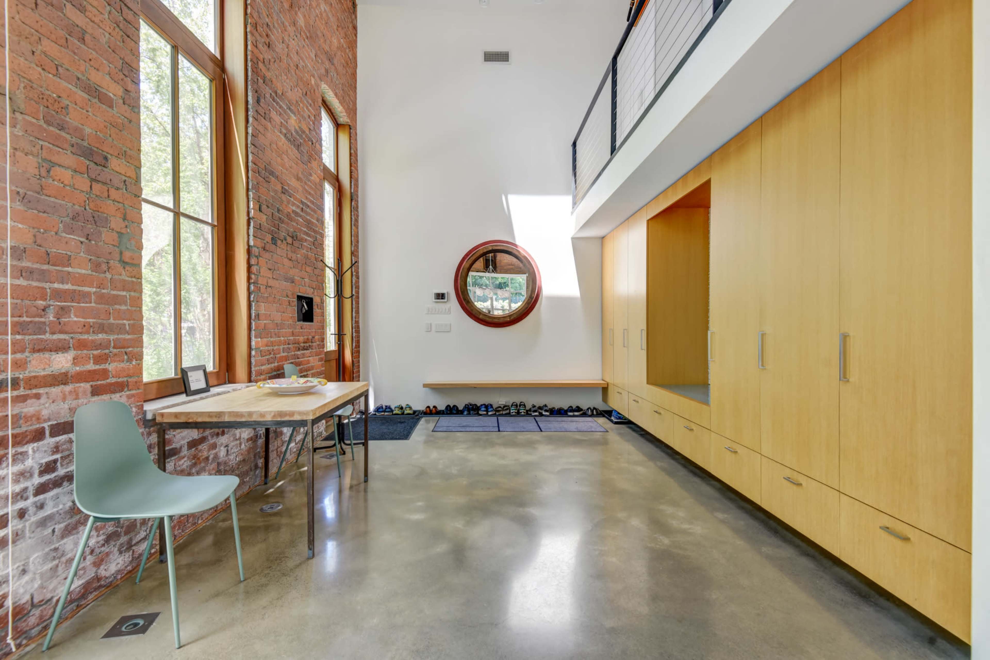 The image shows a modern interior space featuring a wooden table and chair, a round window, and built-in cabinets against a brick wall.
