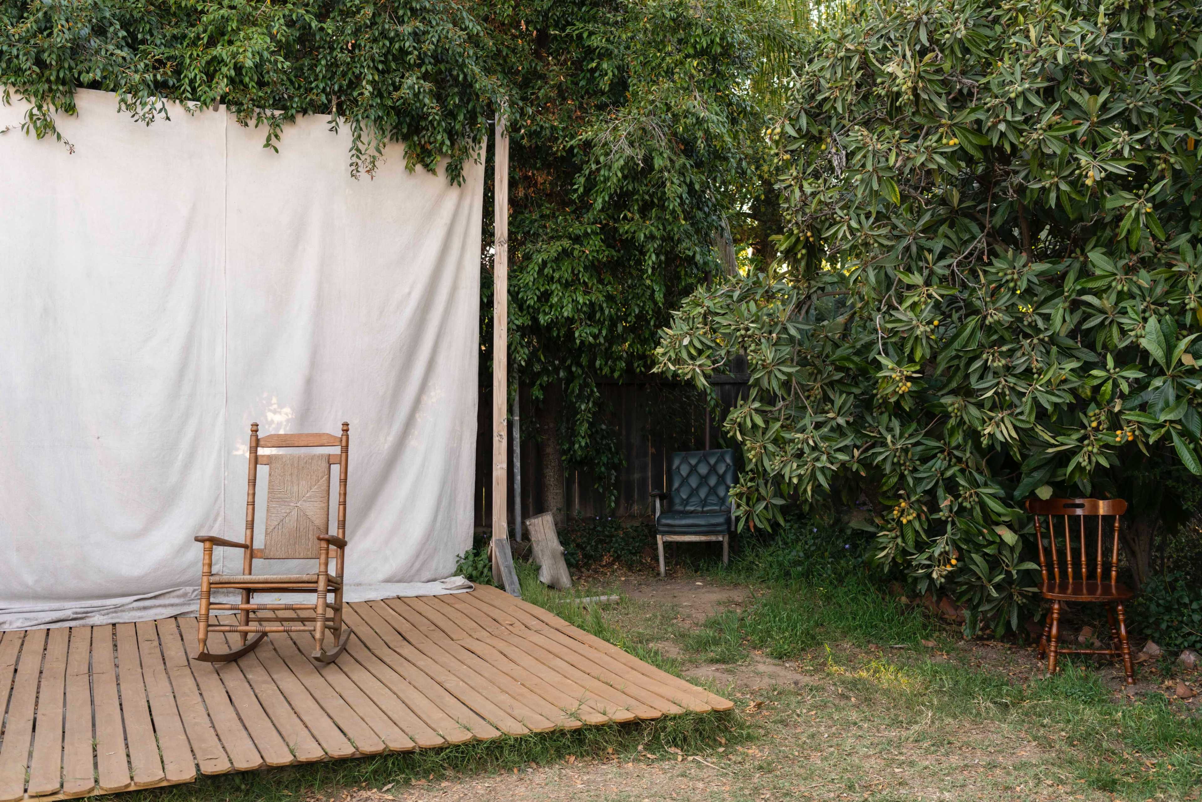 A wooden rocking chair sits on a wooden platform in front of a white backdrop, with two other chairs positioned nearby among greenery.
