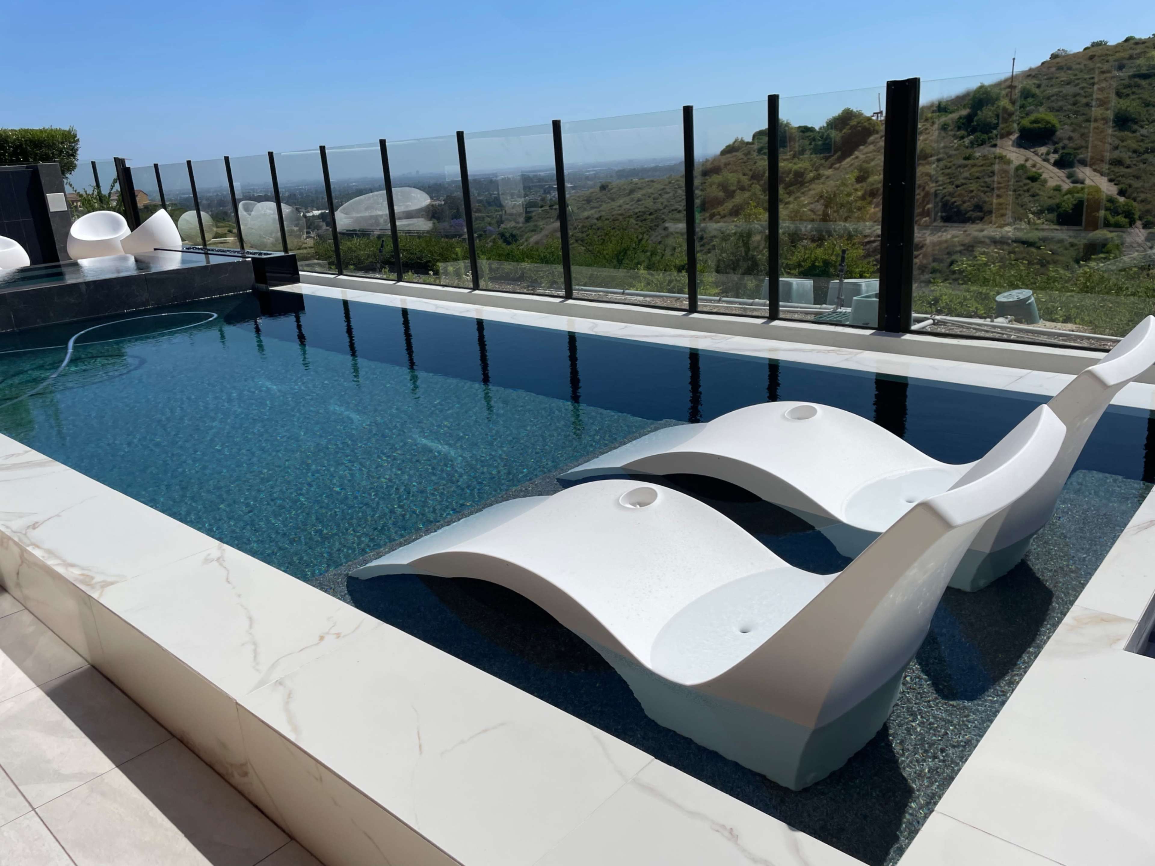 The image shows a modern pool with two white lounge chairs partially submerged in the water, surrounded by a glass barrier and a scenic hillside in the background.