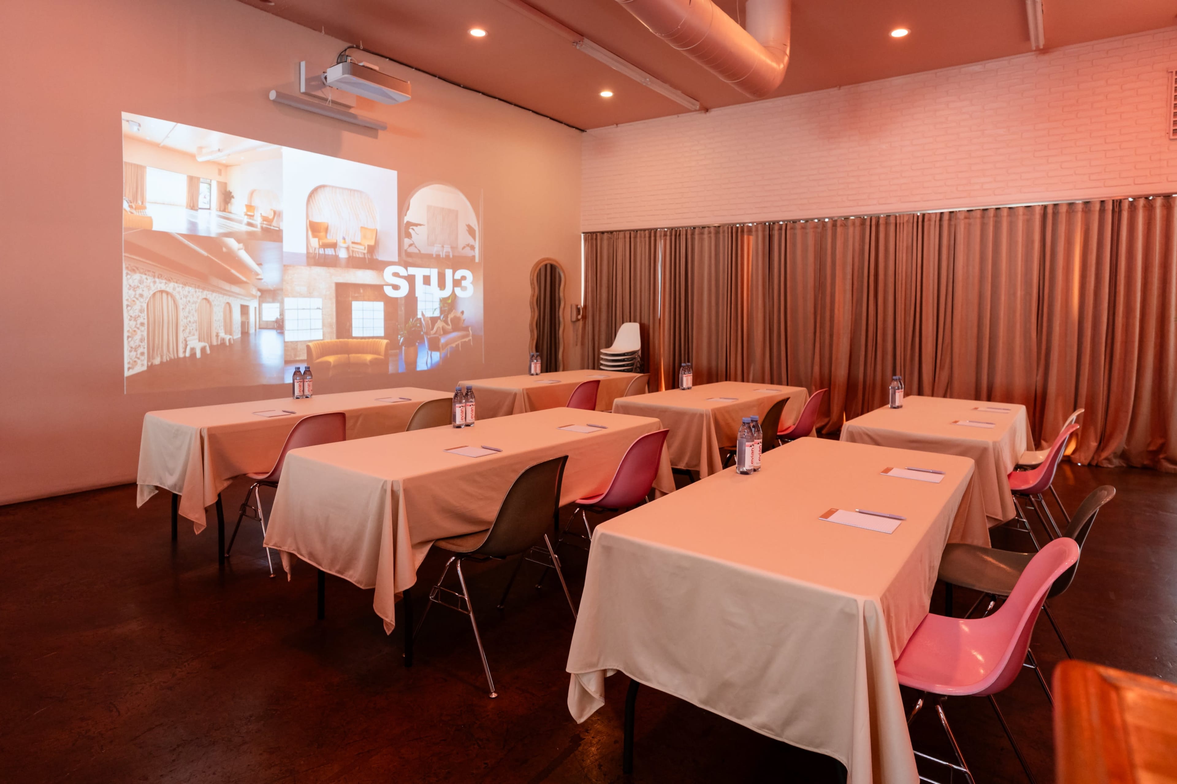 The image shows a meeting room set up with several tables covered in white tablecloths, each featuring a notepad and a water bottle, and a large projector screen displaying images on one wall.