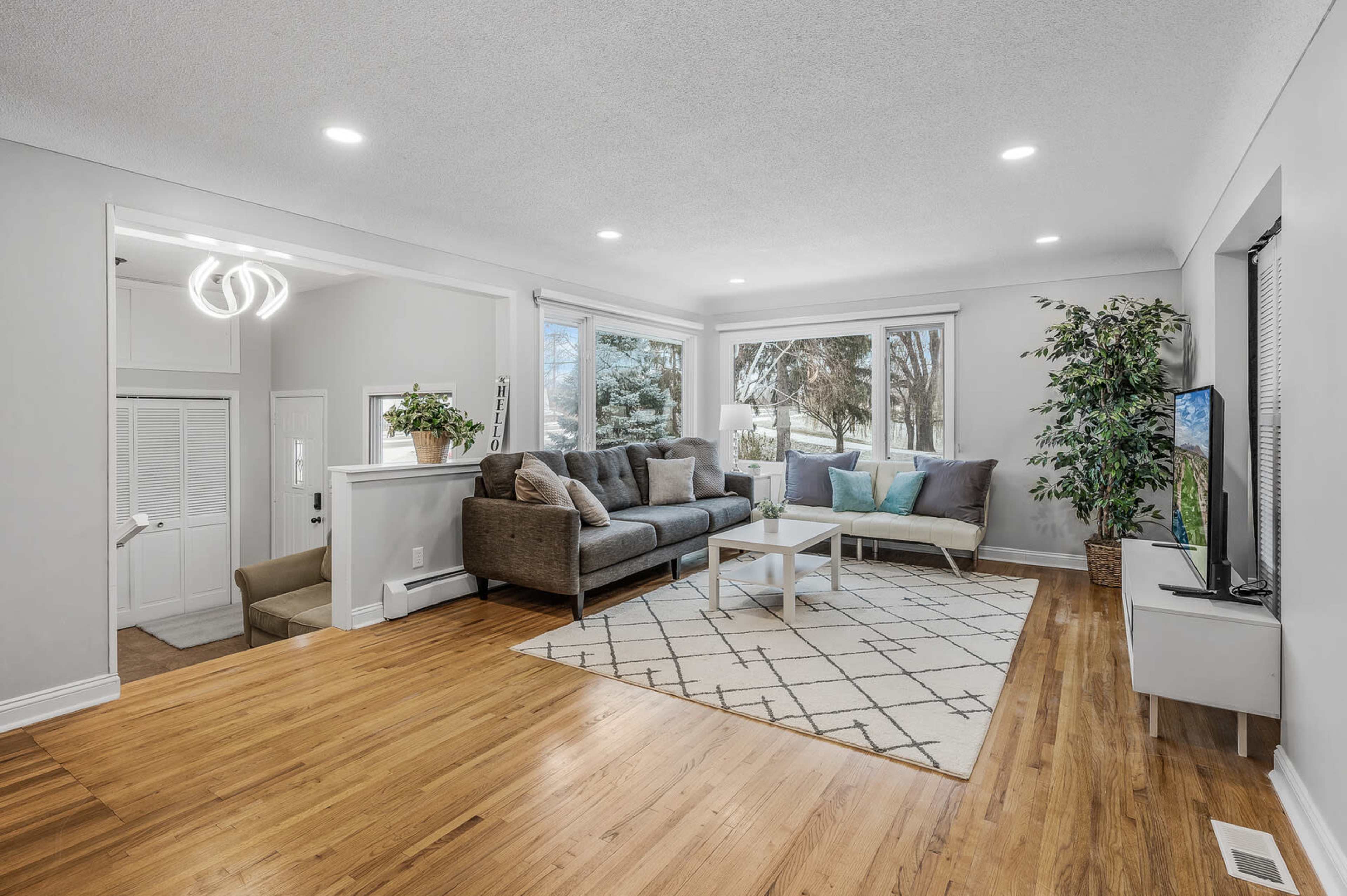 A modern living room features a gray sectional sofa, a light rug with a geometric pattern, and large windows that provide natural light.
