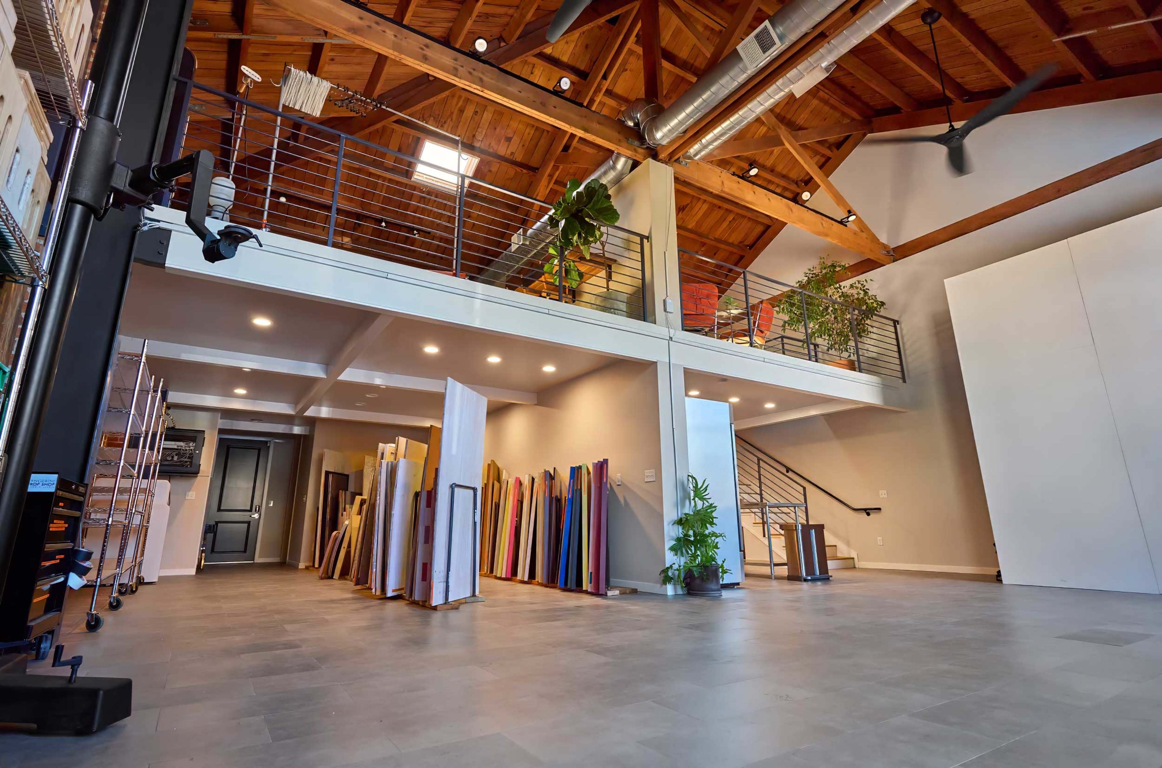 The image shows a spacious interior with high wooden ceilings, featuring a large area for storing colorful materials against a wall and a staircase leading to an upper level.