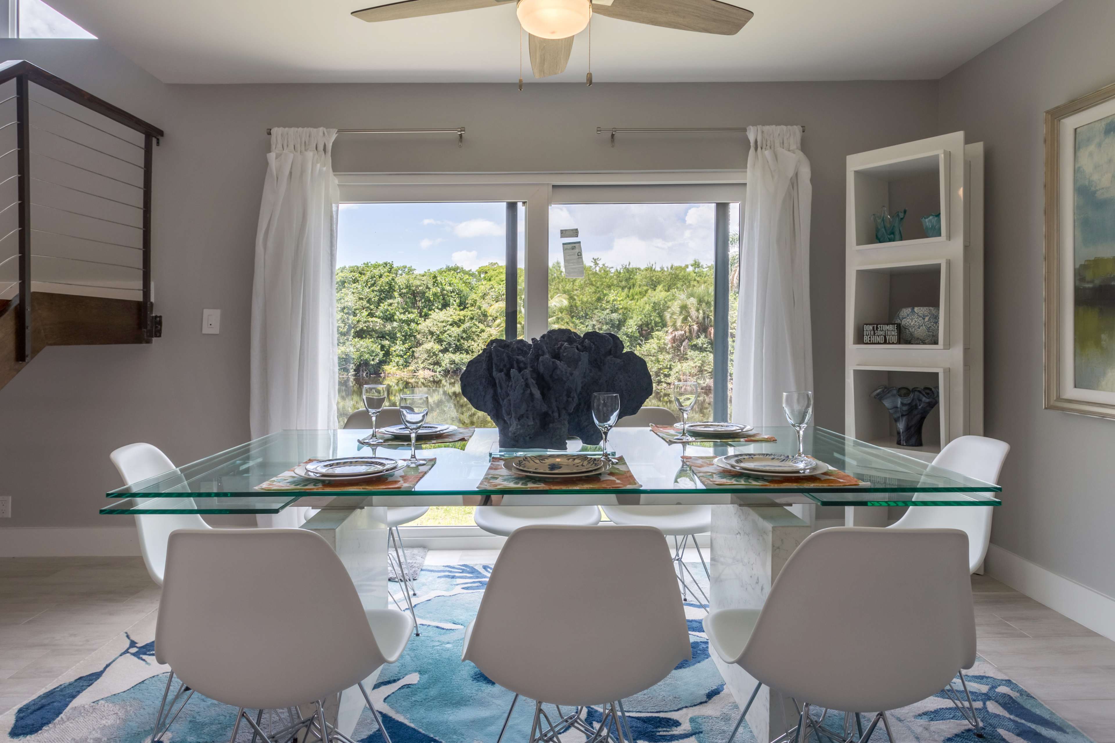 A modern dining room features a glass table set for dinner, surrounded by white chairs, with large windows offering a view of greenery outside.