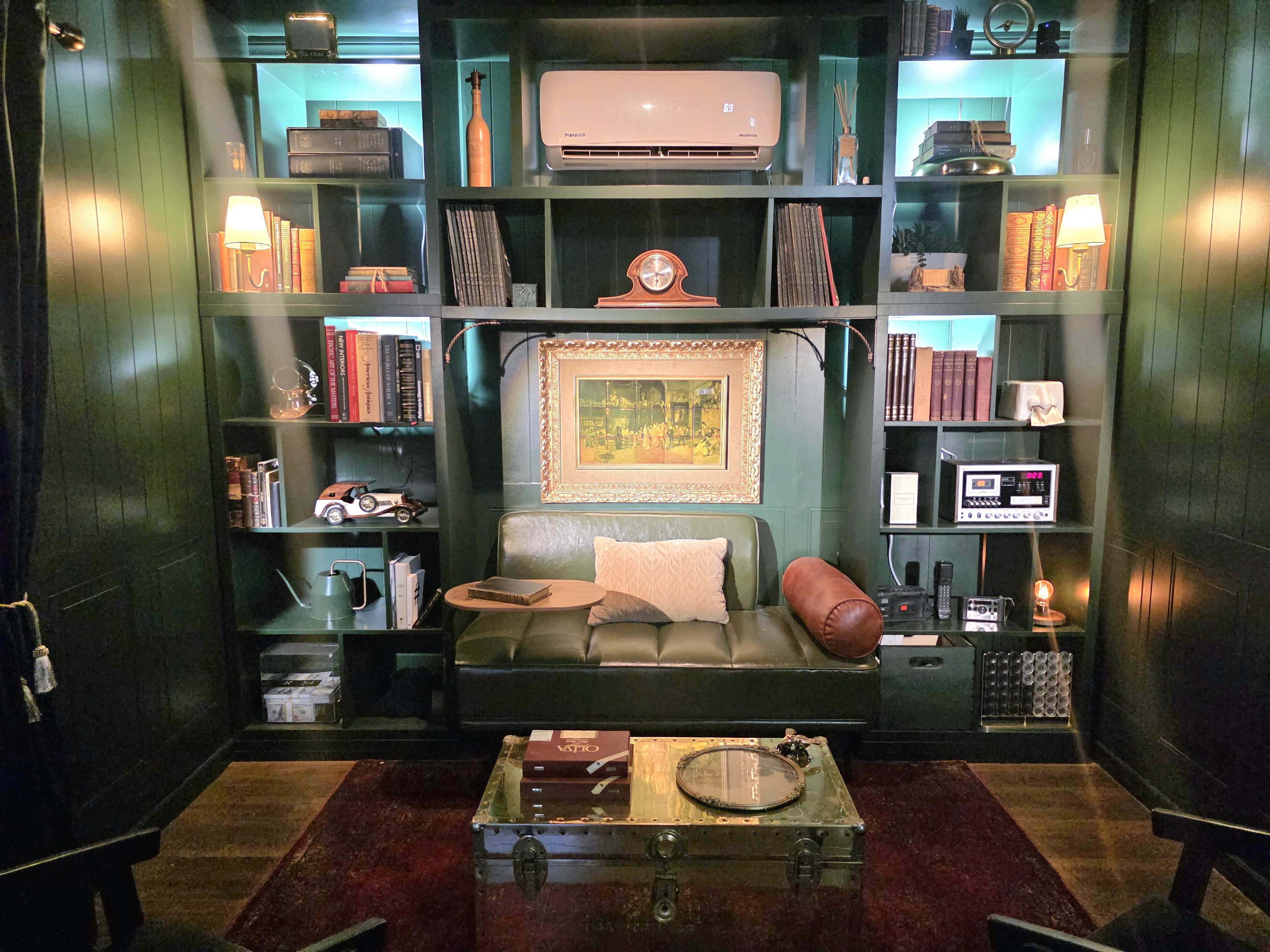 The image shows a cozy reading nook with a green bookshelf, featuring books, decor, and a vintage painting above a leather couch and a wooden coffee table.